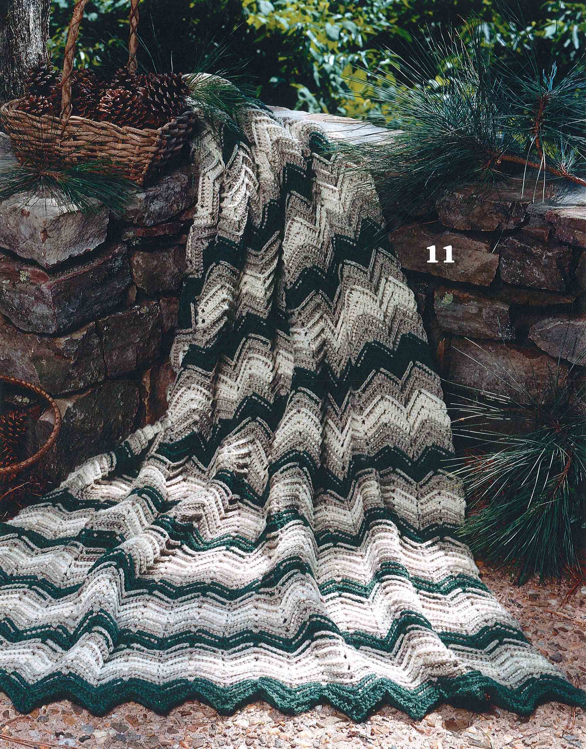 A beginner afghan from Leisure Arts' "Rippling Effects—16 Afghan Designs to Add to Your Home Decor" digital download, made with green, gray, and cream worsted yarn in a ripple pattern, drapes over a stone wall outdoors among pine branches and a wicker basket.