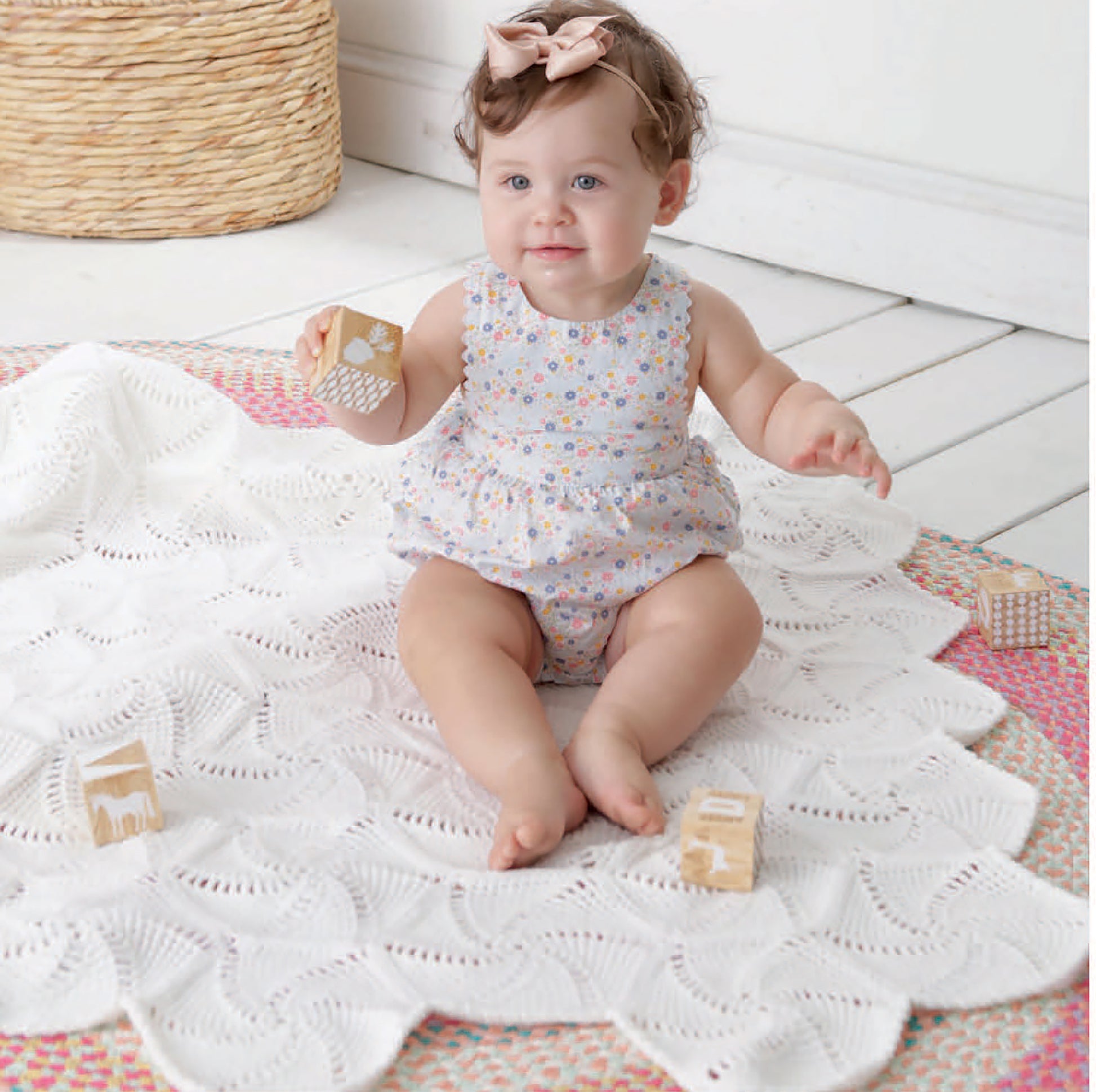 A baby in a floral romper and pink bow sits on the "Our Best Baby Afghans" blanket by Leisure Arts, holding a wooden block. Three more blocks and a woven basket are nearby on the white floor. Digital Download available.