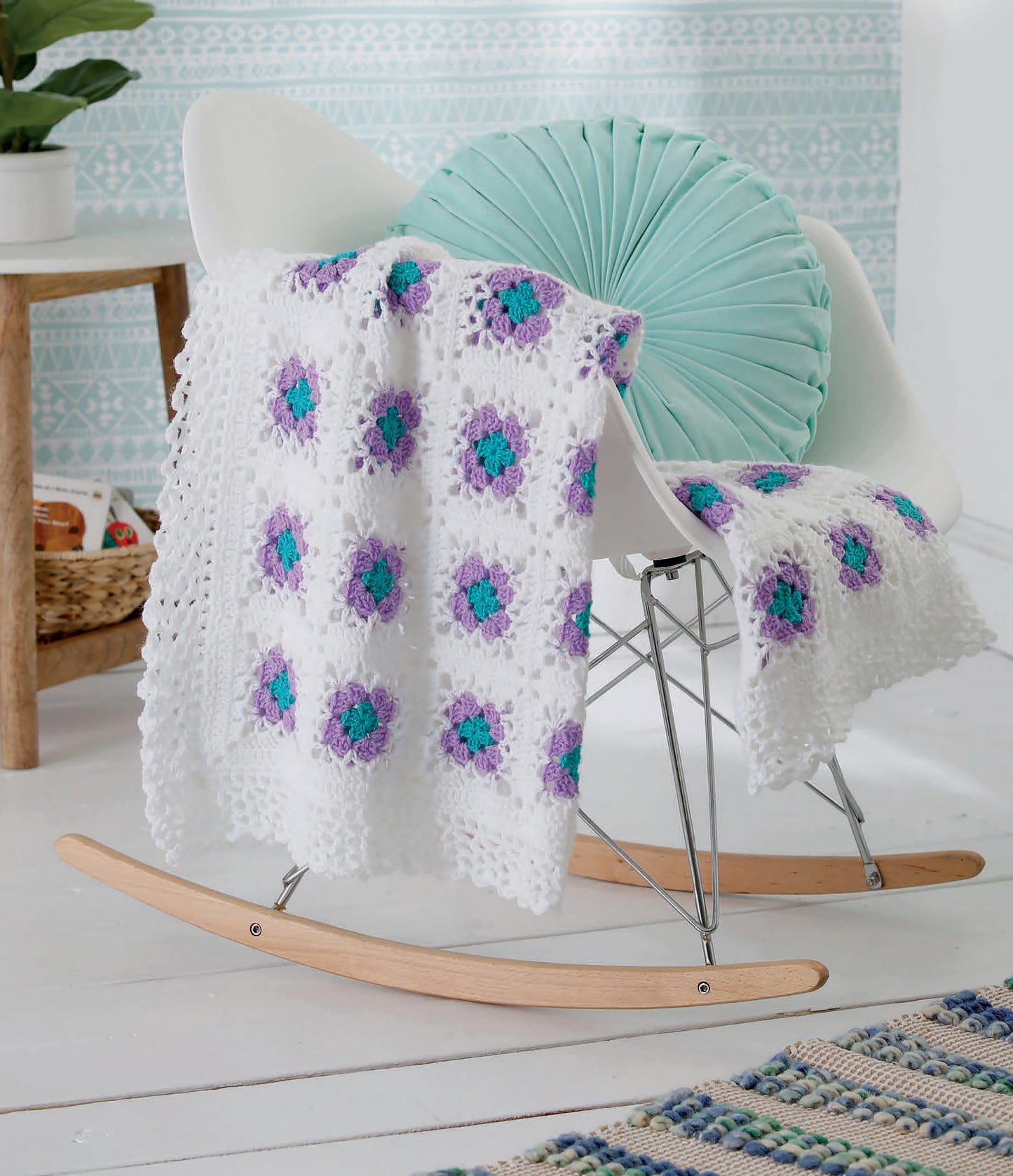 A white rocking chair with wooden rockers displays a mint green cushion and a white crocheted blanket like those in Leisure Arts’ "Our Best Baby Afghans Digital Download," featuring purple and teal flower motifs, set in a bright, cozy room with patterned wallpaper.