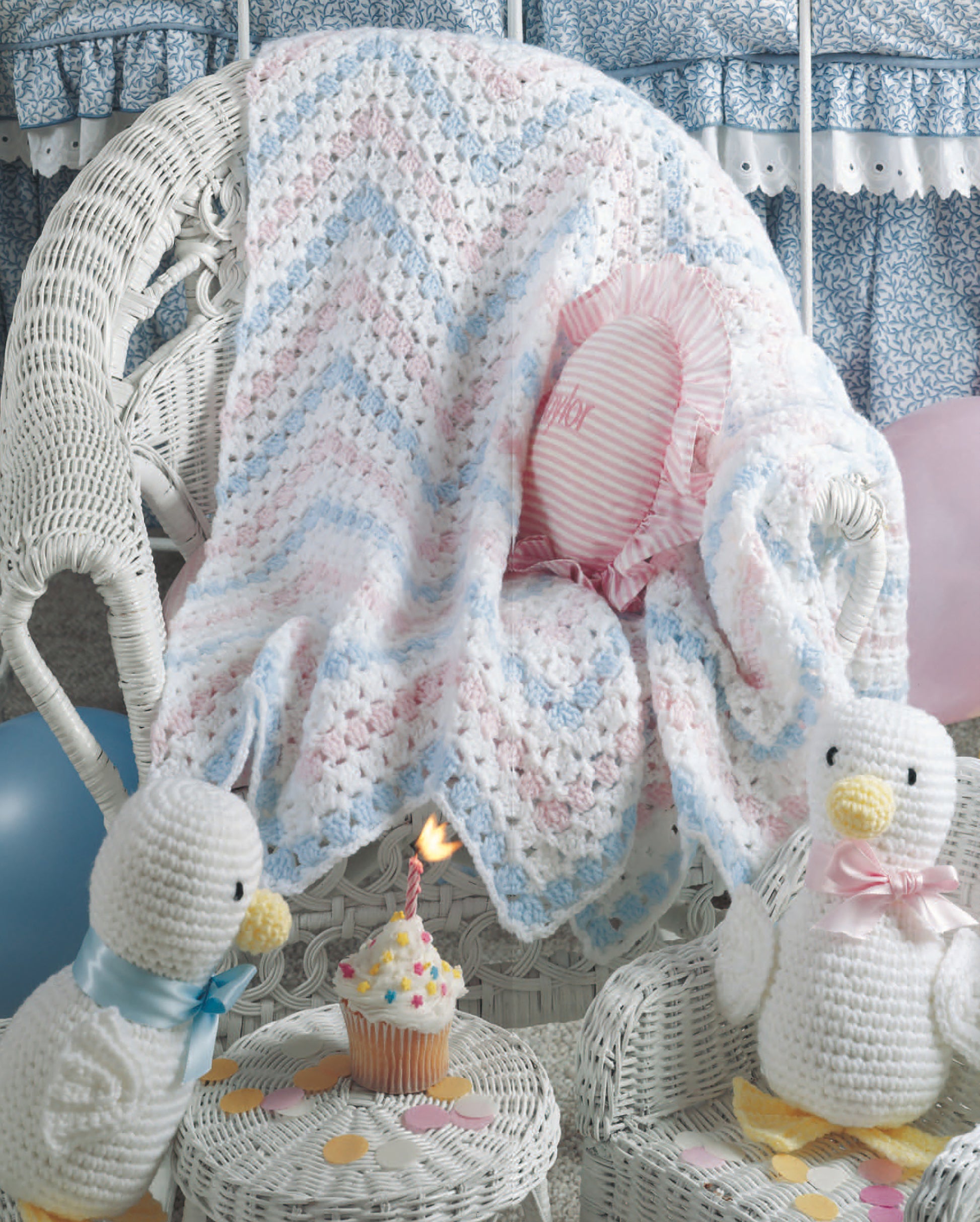 The Leisure Arts Just Ducky Baby Afghans Digital Download is shown draped over a wicker chair, with crocheted duck toys, nursery gifts, a cupcake with candle, balloons, confetti, and a "baby" pillow in a cozy room.