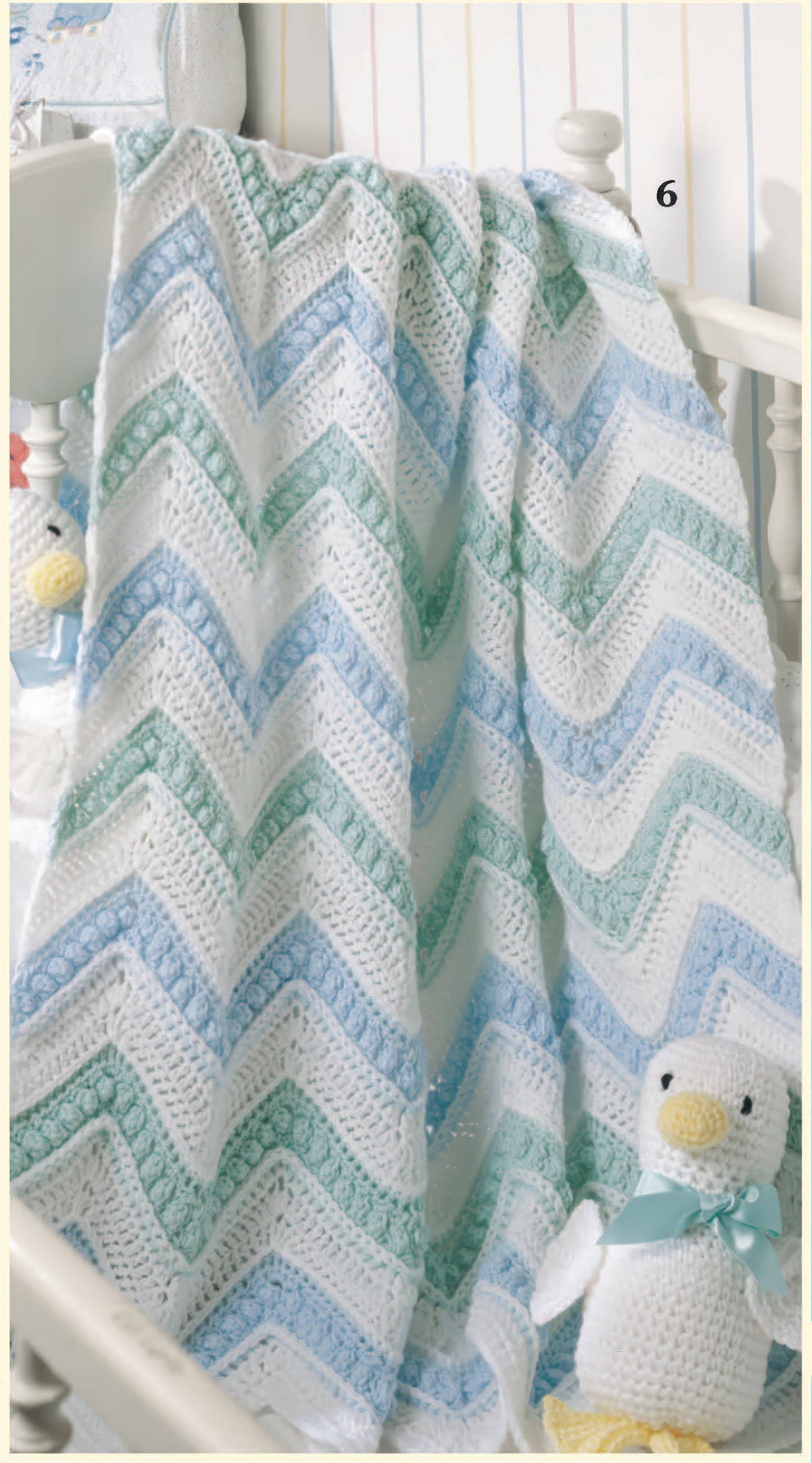 The Just Ducky Baby Afghans Digital Download by Leisure Arts features a crocheted zigzag blanket in white, green, and blue draped over a crib rail, paired with a plush duck toy with a blue ribbon—an adorable nursery gift.