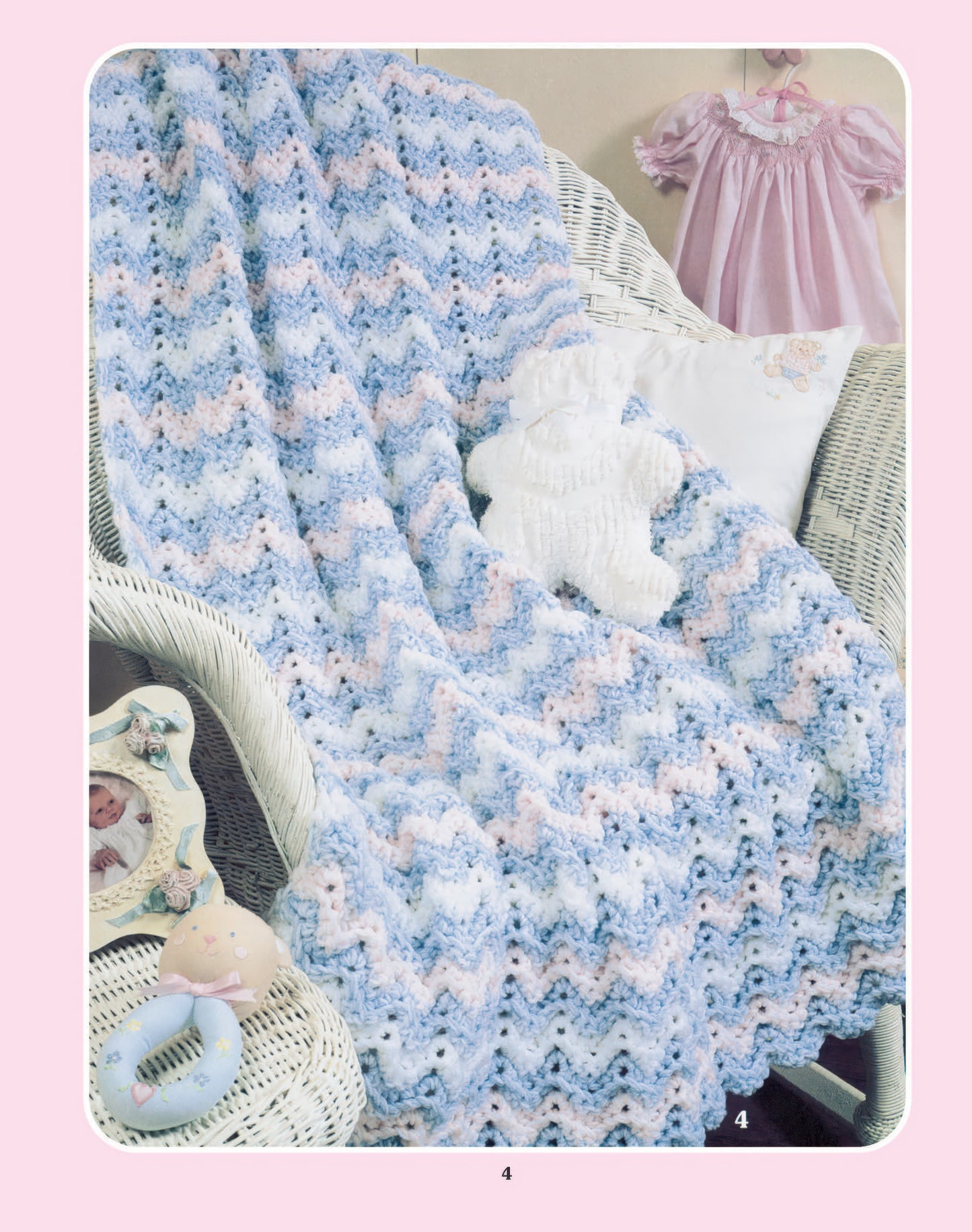 A cozy nursery features the "More Double-Quick for Baby" digital download by Leisure Arts: a pastel zigzag crocheted blanket (worsted yarn) drapes on a white wicker chair, with a teddy bear, pink baby dress, pillow, and rattle nearby.
