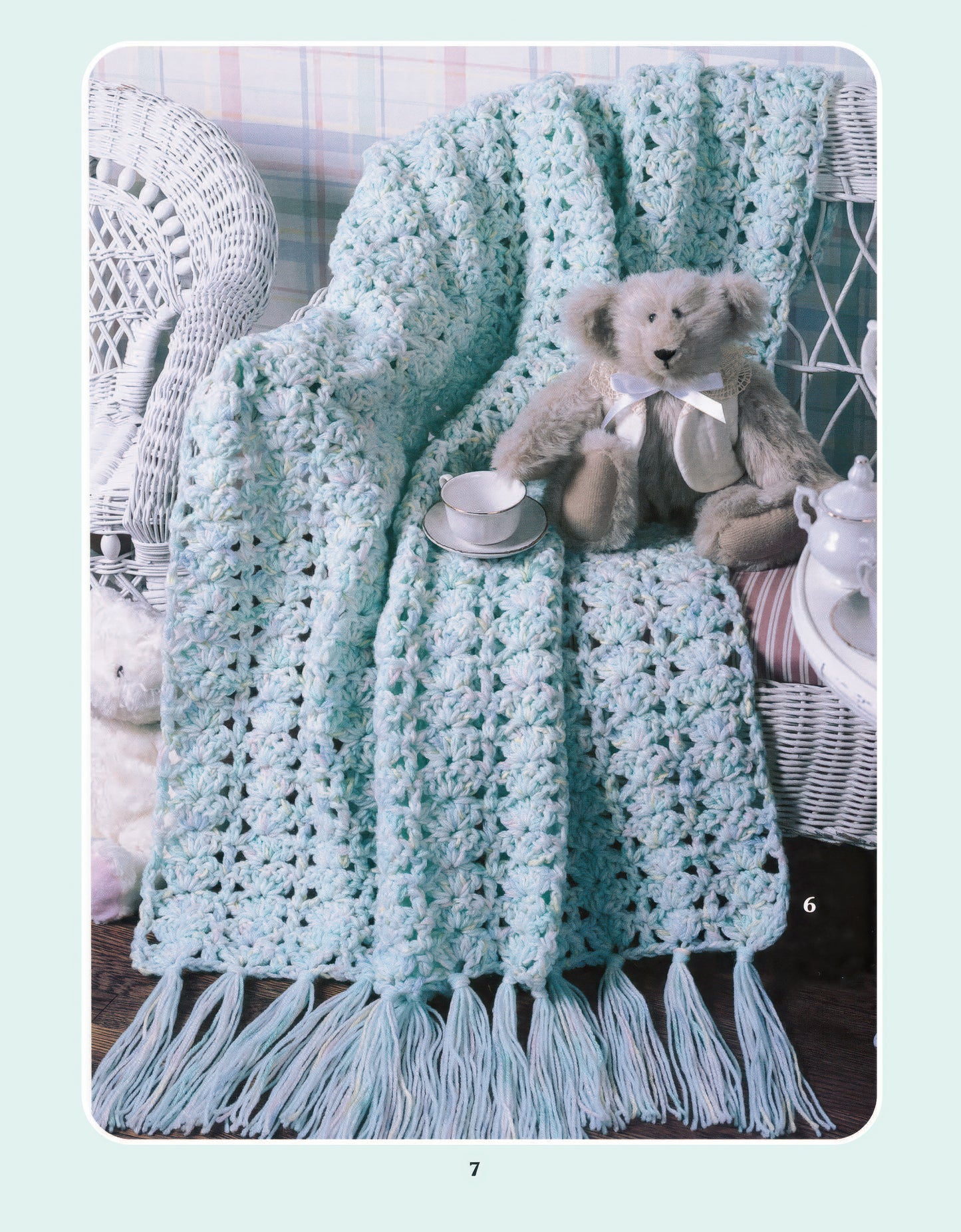 The More Double-Quick for Baby Digital Download by Leisure Arts showcases a light blue worsted weight crochet blanket draped on a white wicker chair, beside a teddy bear and teacup, set against a pastel plaid—ideal for beginner crocheters.