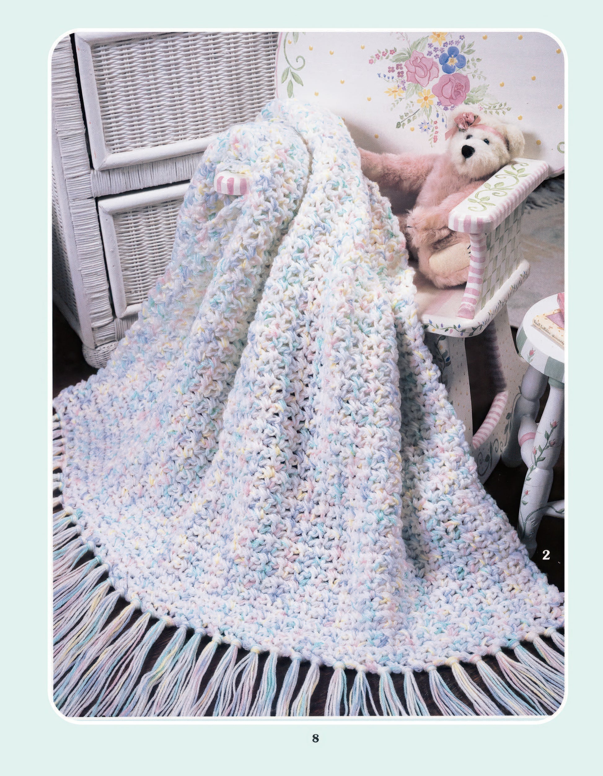 The More Double-Quick for Baby Digital Download by Leisure Arts features a pastel crocheted blanket with fringe, made from worsted yarn, draped over a white wicker chair beside a teddy bear and a floral-painted rocker.