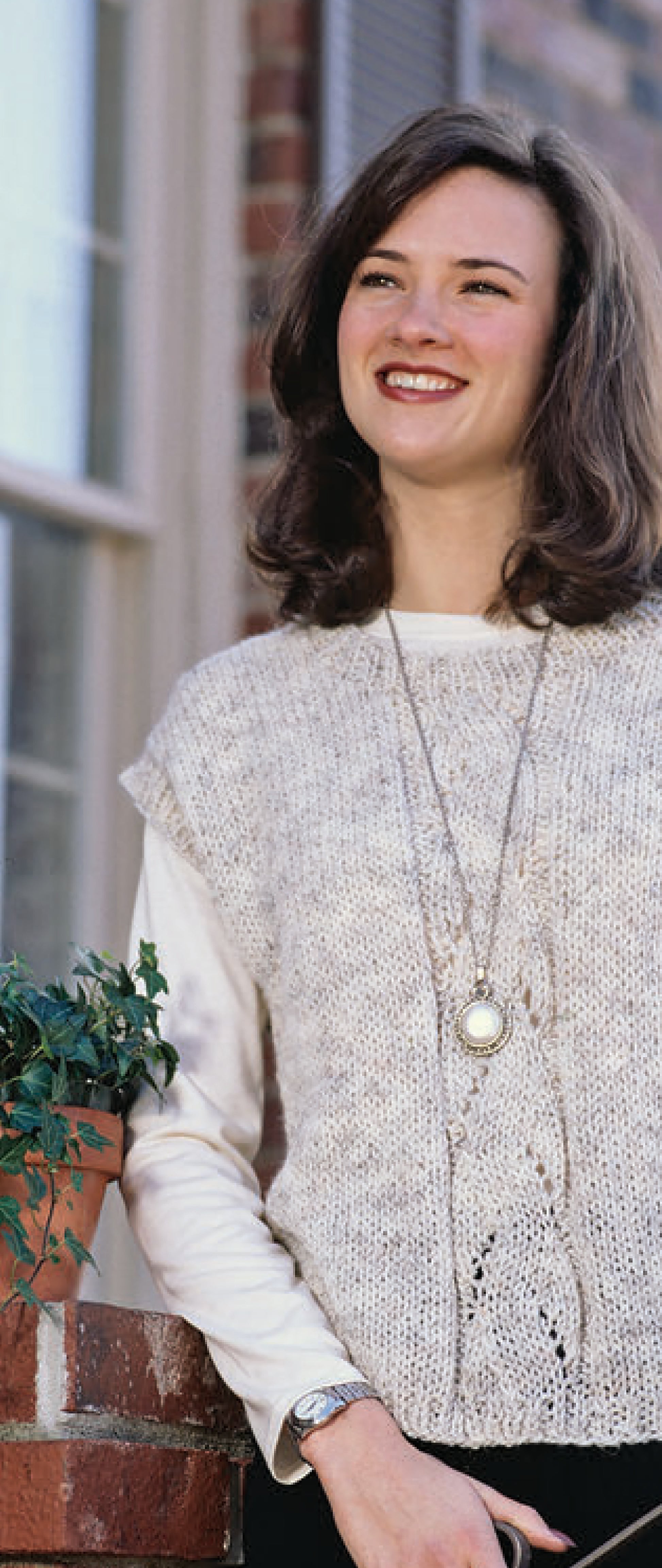 A woman with shoulder-length brown hair smiles beside a potted plant outdoors, wearing a cream knit sweater made from patterns in Leisure Arts’ The Big Book of Quick Knit Sweaters Digital Download. A building and window are visible in the background.