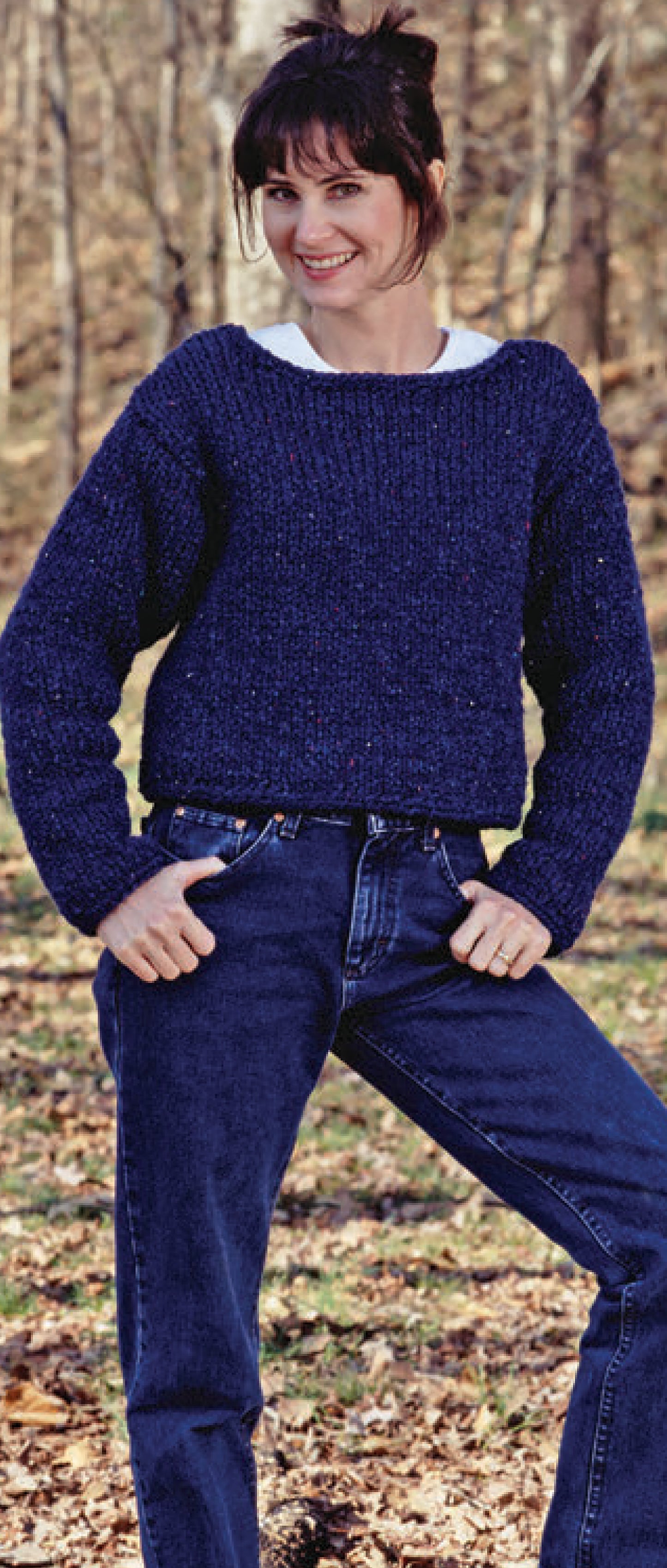 A woman with dark hair in a bun smiles outdoors among trees and fallen leaves. She wears a navy blue sweater inspired by classic designs featured in "The Big Book of Quick Knit Sweaters" digital download from Leisure Arts.