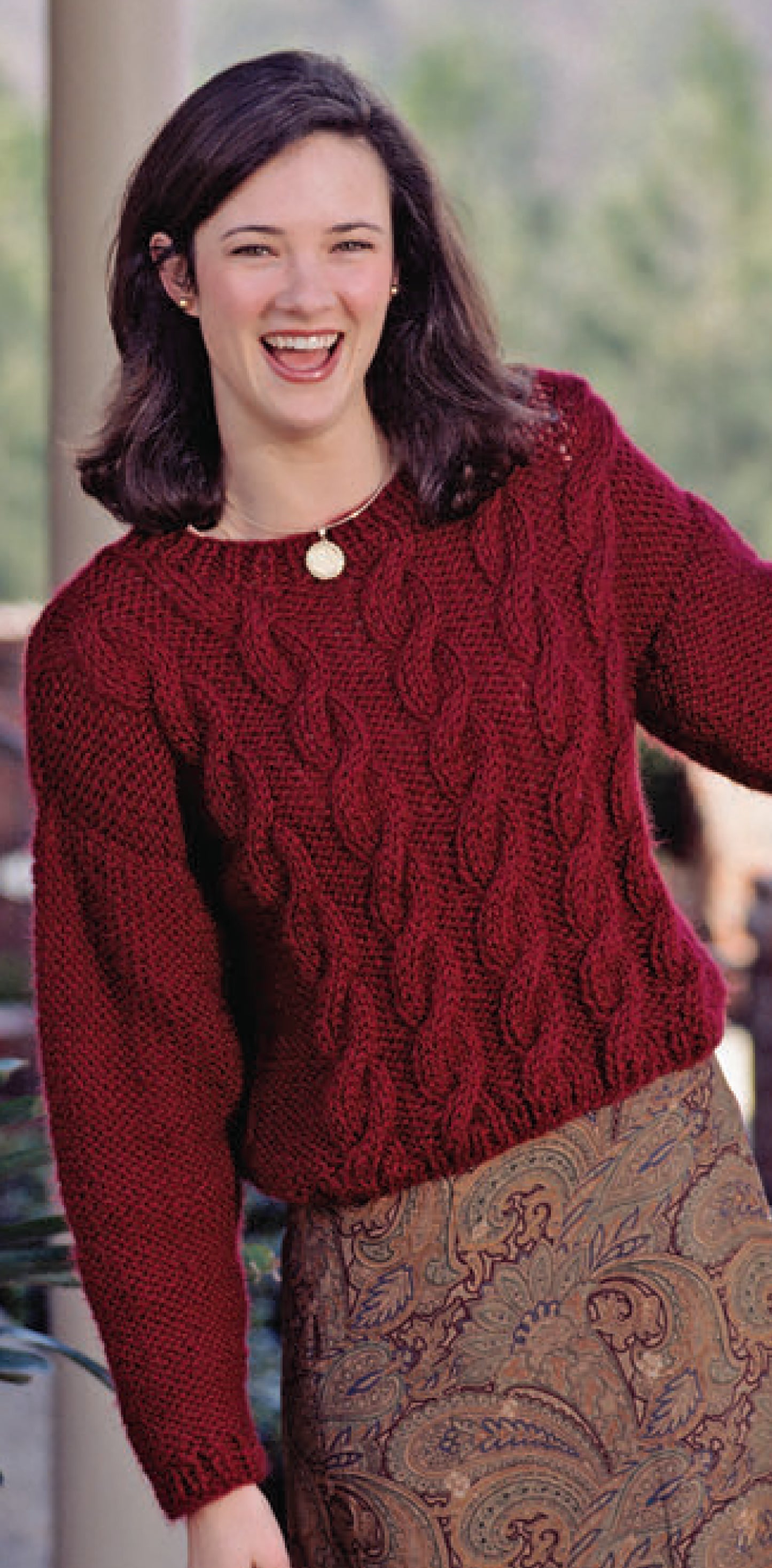 A woman with shoulder-length dark hair smiles while wearing a sweater made from "The Big Book of Quick Knit Sweaters" digital download by Leisure Arts, standing outdoors with greenery blurred in the background.