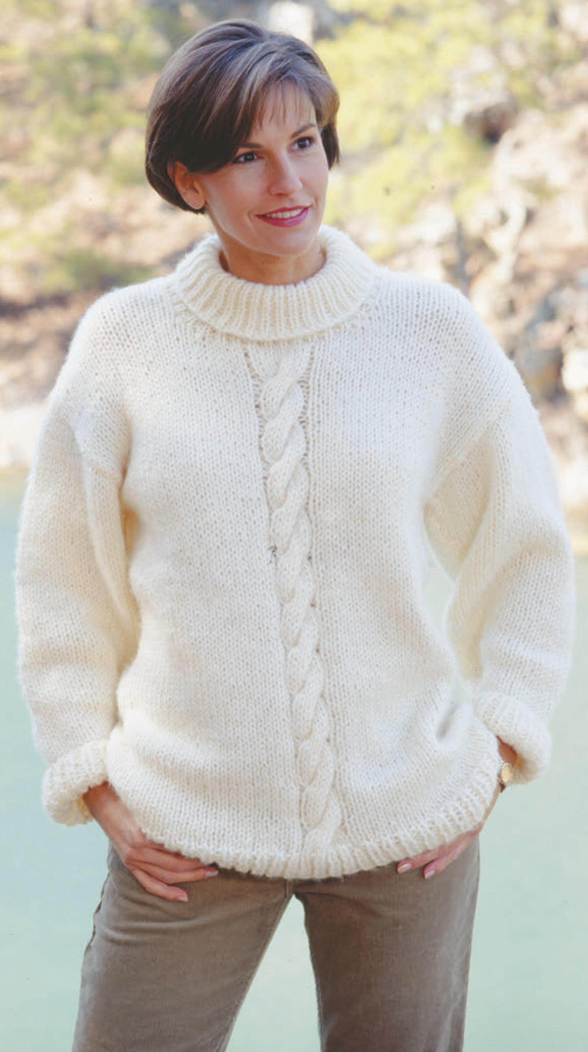 A woman smiles outdoors in a cozy, cream cable-knit turtleneck sweater, as featured in "The Big Book of Quick Knit Sweaters" by Leisure Arts. She pairs it with brown pants; blurred trees and water complete the scenic background.