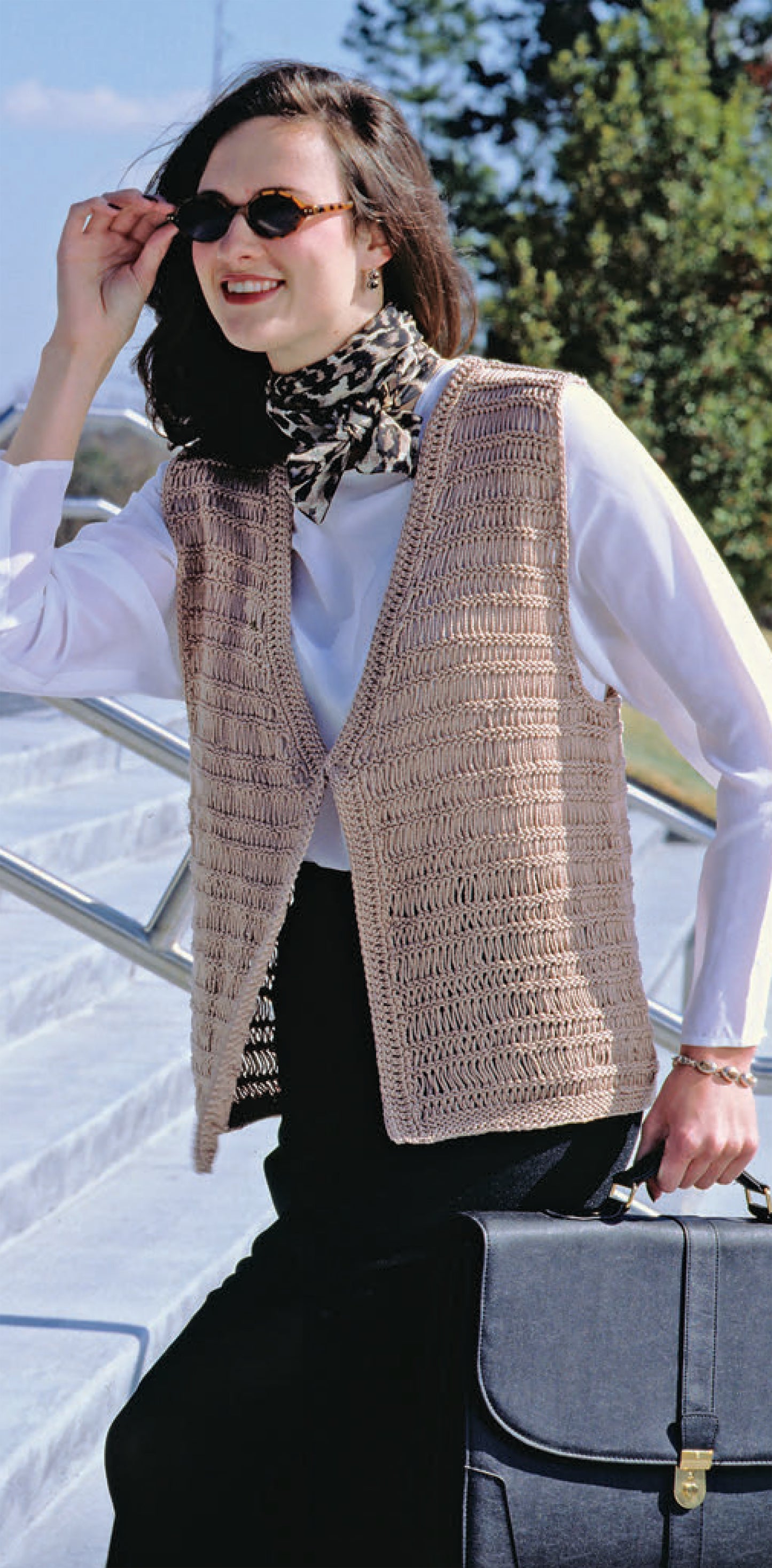 A woman outdoors in sunglasses, a white blouse, black skirt, and a beige vest modeled from Leisure Arts’ The Big Book of Quick Knit Sweaters Digital Download stands on steps, holding a scarf and carrying a black briefcase with trees and blue sky behind her.
