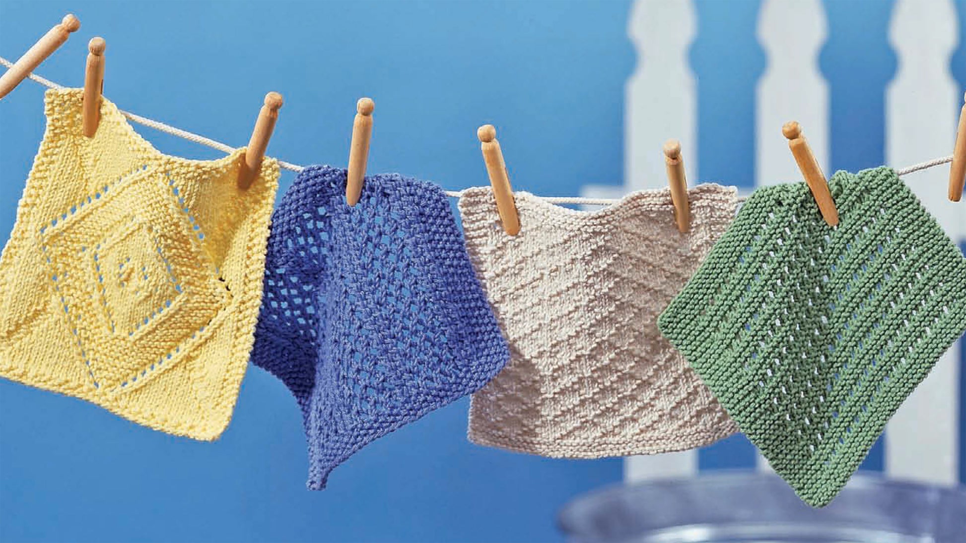 Four colorful cotton dishcloths in yellow, blue, beige, and green from the Nifty Knit Dishcloths Digital Download by Leisure Arts hang from a clothesline. Beginner-friendly patterns feature unique textures, perfect for easy knitting projects.