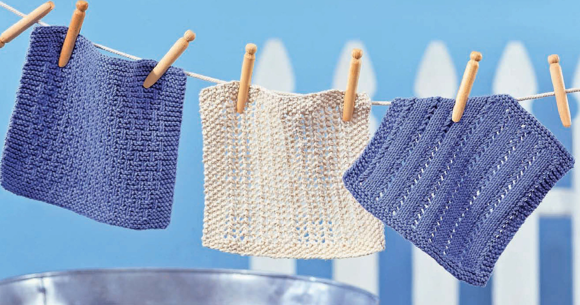 Three Nifty Knit Dishcloths from Leisure Arts—two blue, one beige—hang with wooden clothespins against a blue background and white picket fence, highlighting beginner-friendly digital patterns available for download.