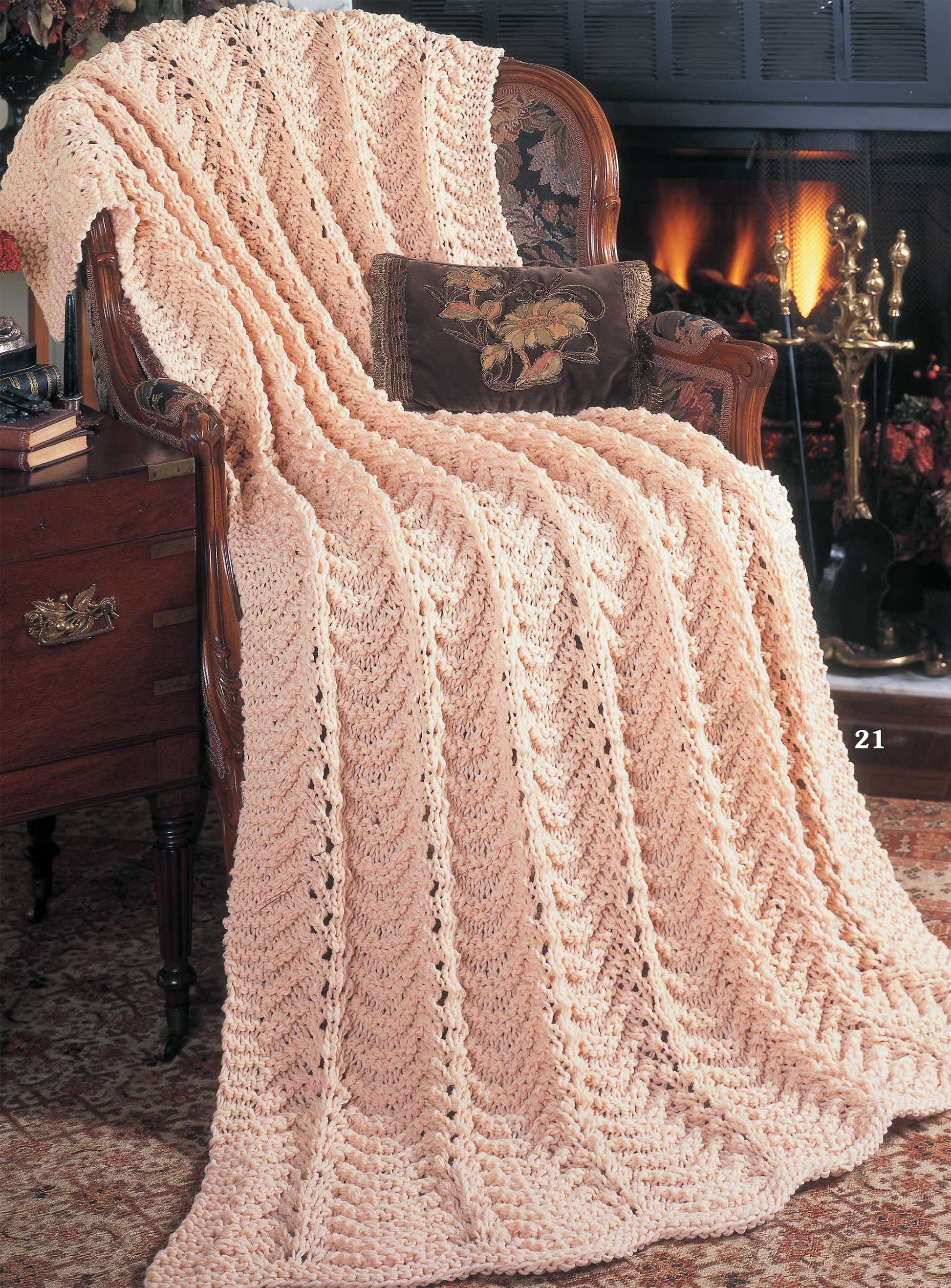 A cozy knit blanket, made with Leisure Arts’ Big Book of Quick Knit Afghans Digital Download, drapes over an ornate wooden chair beside a side table and in front of a glowing fireplace adorned with brass fire tools.