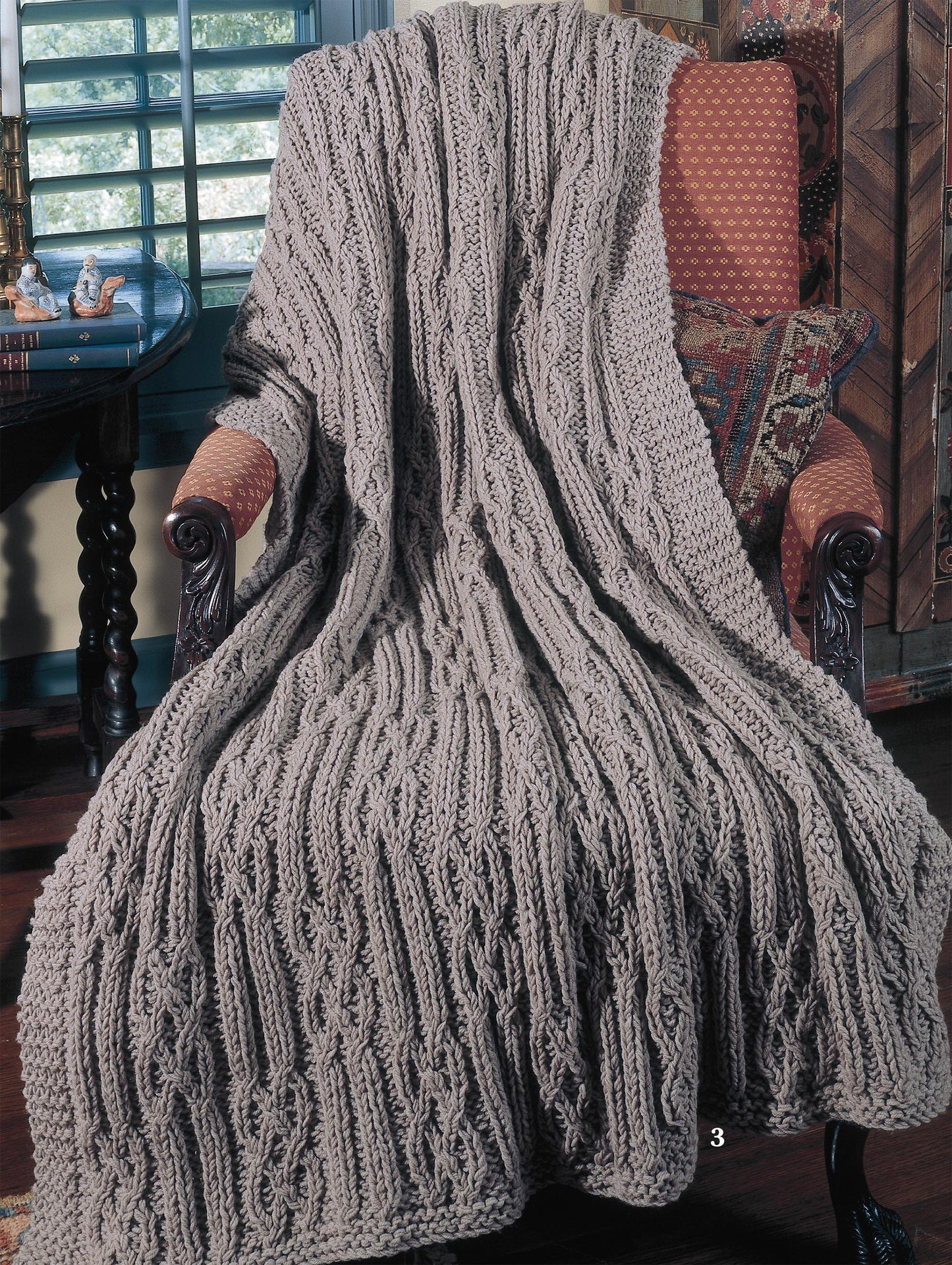 A cozy room features a light brown cable-patterned afghan from the "Big Book of Quick Knit Afghans" by Leisure Arts, draped over an armchair with patterned decor and plantation shutters, inviting warmth and comfort.