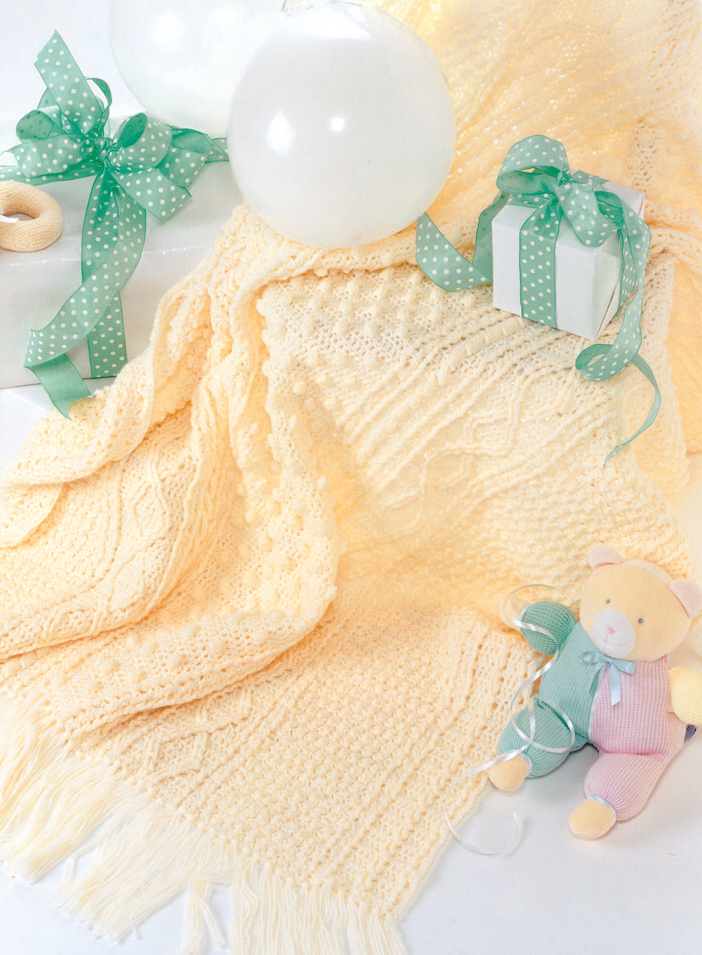 The Snuggle-Up Baby Afghans—8 Projects Digital Download by Leisure Arts is featured with a pale yellow knitted blanket, white balloons, a polka-dot gift box, and a pastel teddy bear—perfect inspiration for beginner crochet fans.