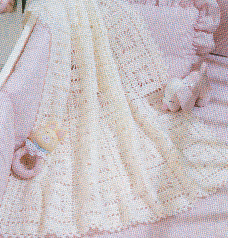 A light pink crib with ruffled bedding displays a delicate white crochet blanket made from Leisure Arts' "Snuggle-Up Baby Afghans—8 Projects" digital download. Plush toys, like a cat rattle and pink lamb, complete this sweet beginner crochet scene.