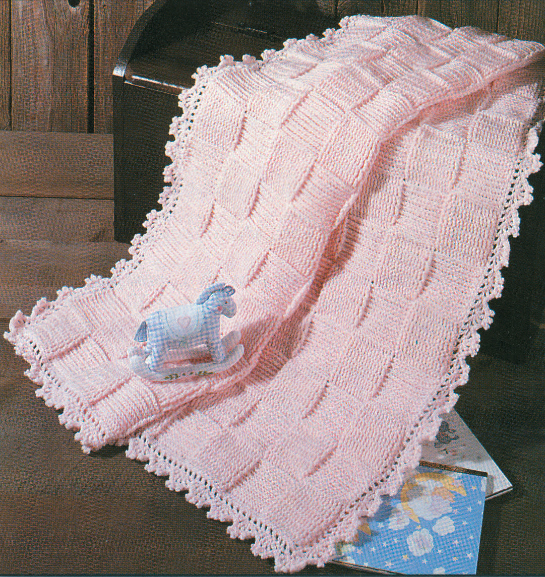 The Leisure Arts Snuggle-Up Baby Afghans—8 Projects digital download features classic crochet patterns like a soft pink checkered blanket with lacy edging, shown draped over a dark chest beside a blue rocking horse and children’s books.