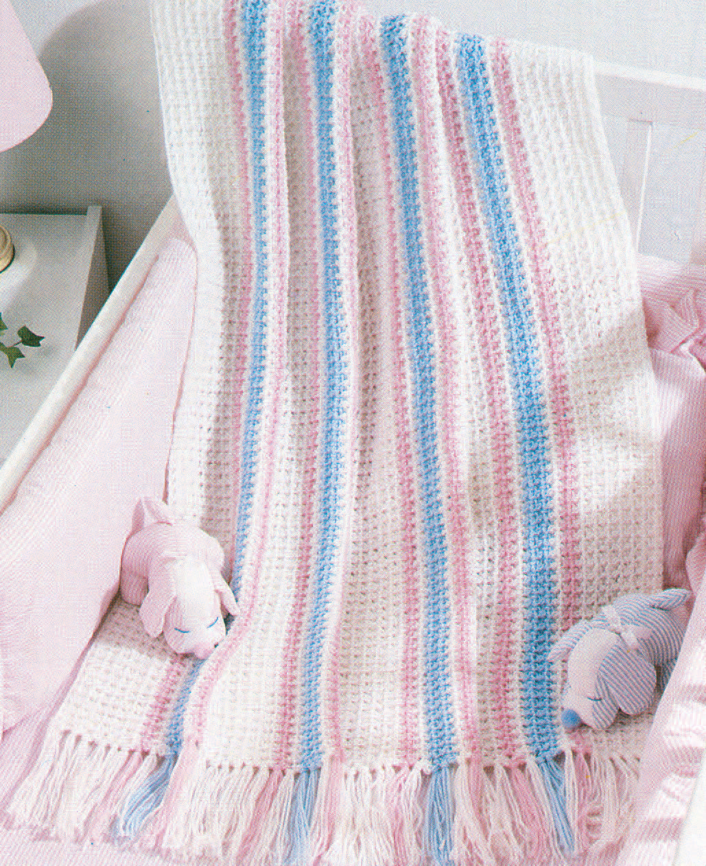 Snuggle-Up Baby Afghans—8 Projects Digital Download by Leisure Arts displays classic white, pink, and blue stripes with tasseled fringes. Draped over a crib between two plush bunnies, it adds charm to soft pink nursery décor.