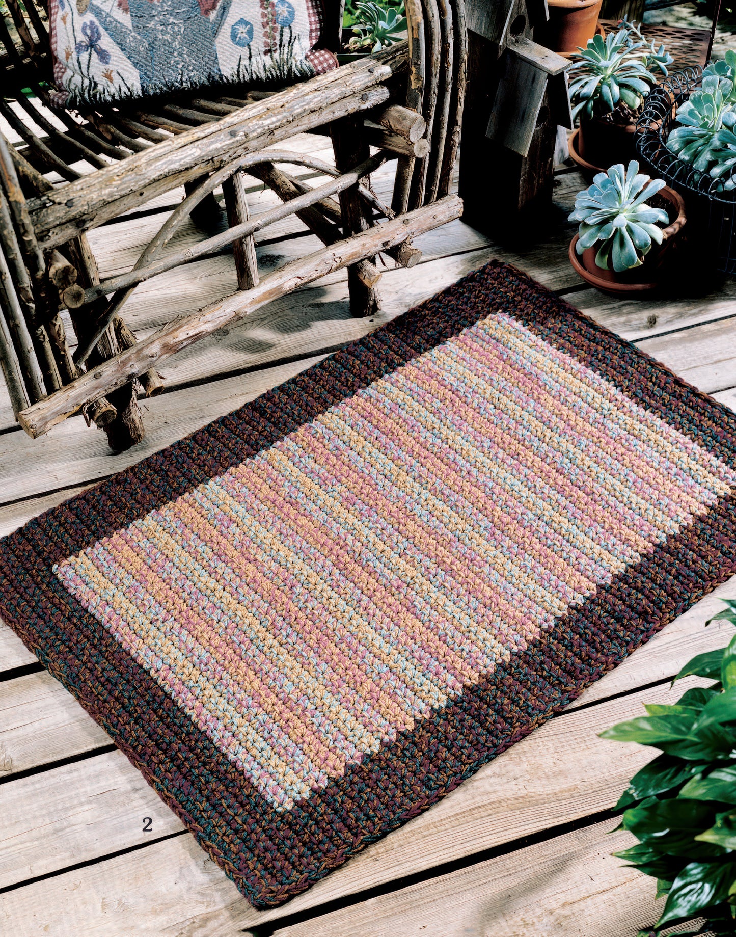 The Easy Rugs to Crochet Digital Download by Leisure Arts features rectangular, multicolored braided rug patterns with dark borders, perfect for adding style to any wooden deck or rustic space with potted plants.