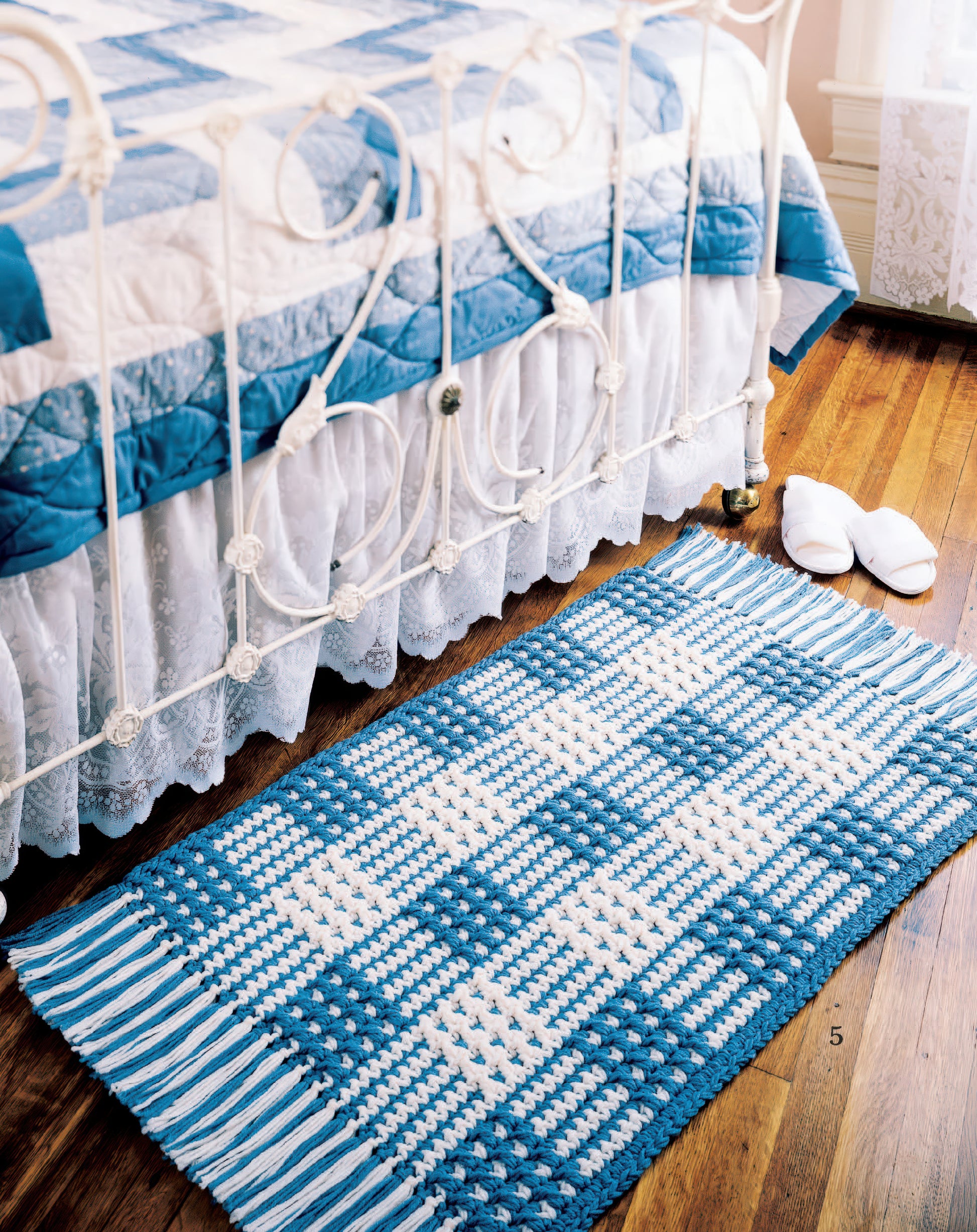 A blue and white checkered crocheted rug from the "Easy Rugs to Crochet Digital Download" by Leisure Arts lies on a wooden floor beside a white metal bed with a matching quilt; white slippers rest nearby.