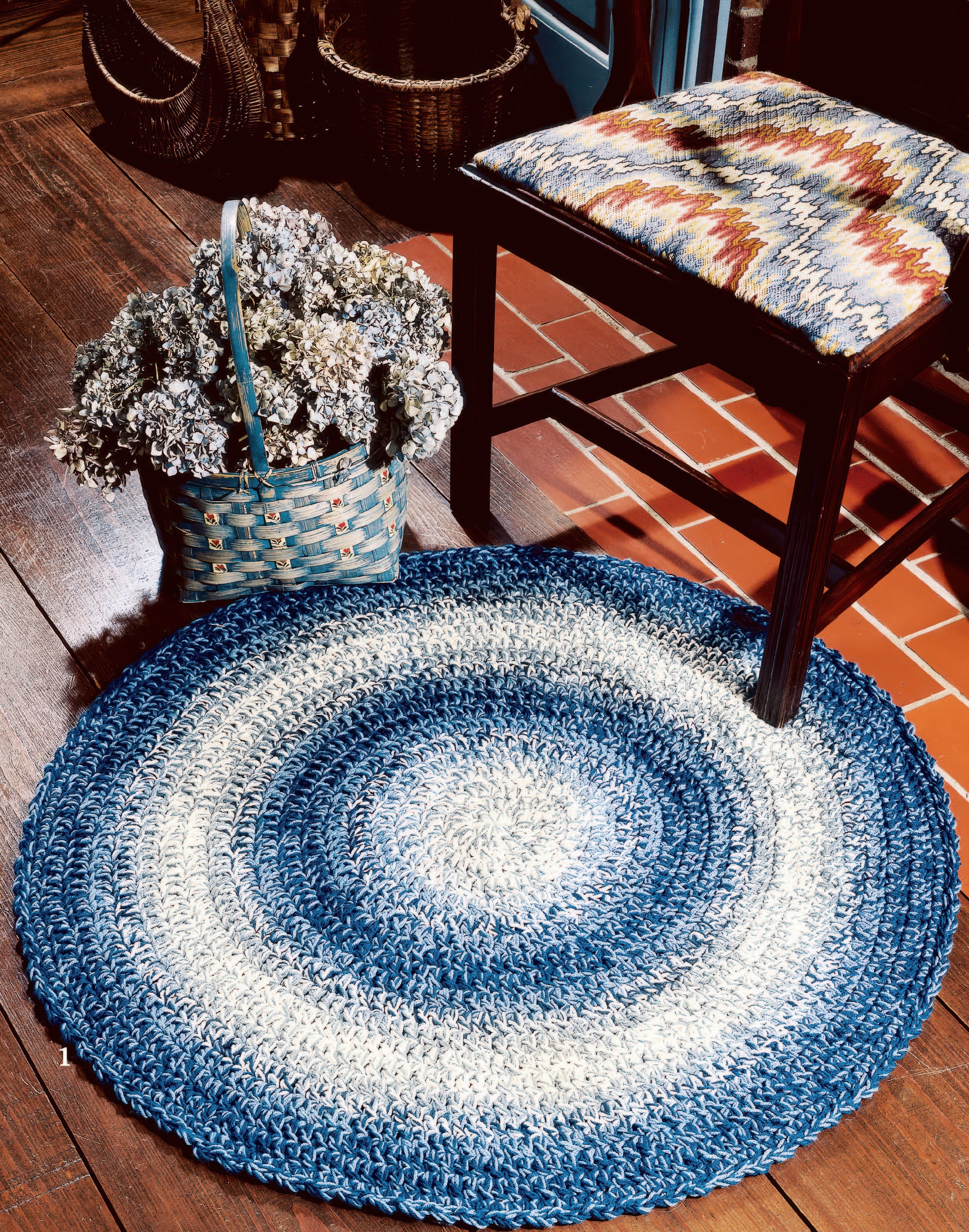 The Easy Rugs to Crochet Digital Download by Leisure Arts features a round blue and white braided rug, perfect for wooden floors, styled here beside a colorful woven chair and a basket of dried flowers.