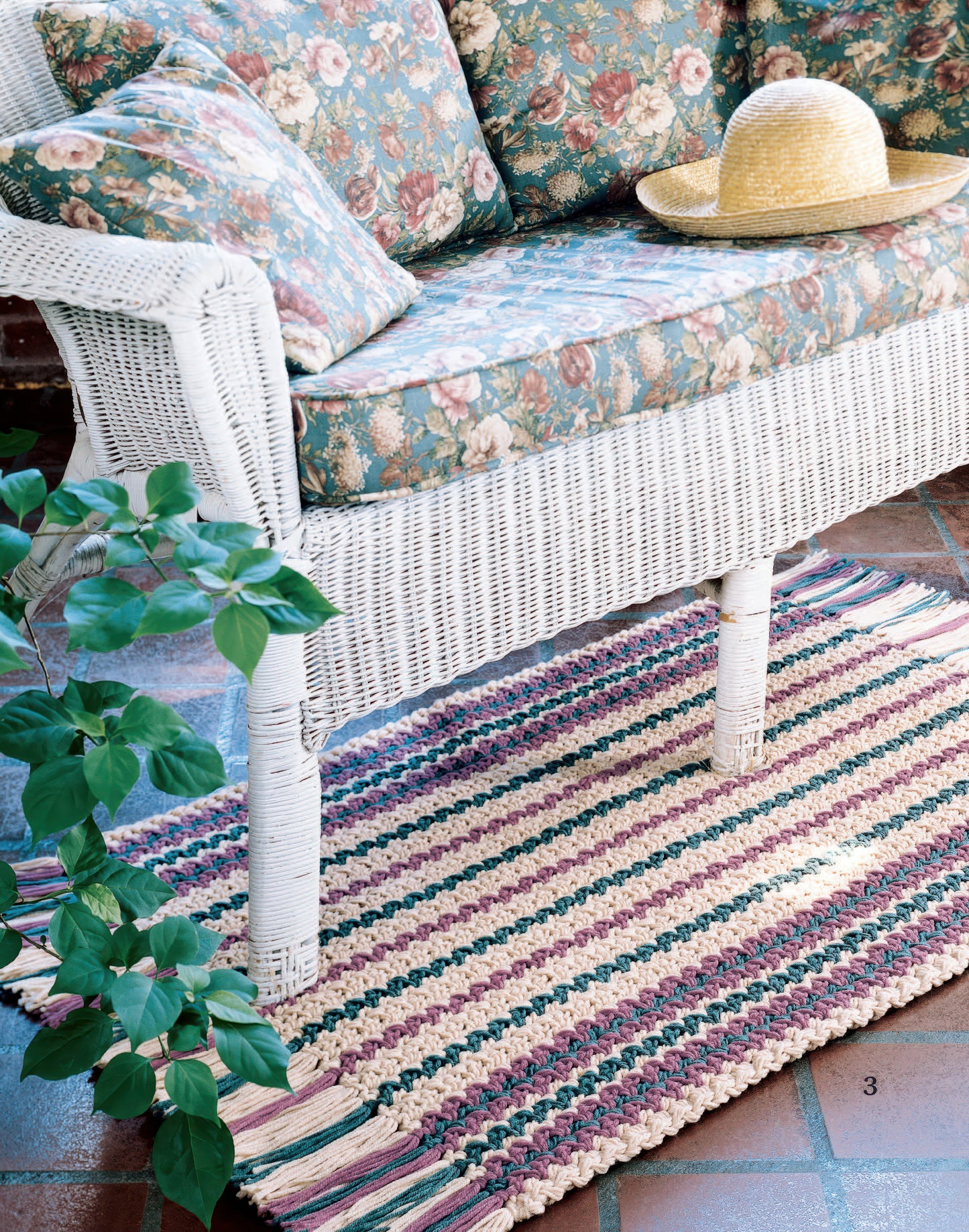 A white wicker loveseat with floral cushions sits on a colorful striped rug with rectangle patterns, crocheted from the "Easy Rugs to Crochet Digital Download" by Leisure Arts, next to a leafy plant and terracotta floor tiles.