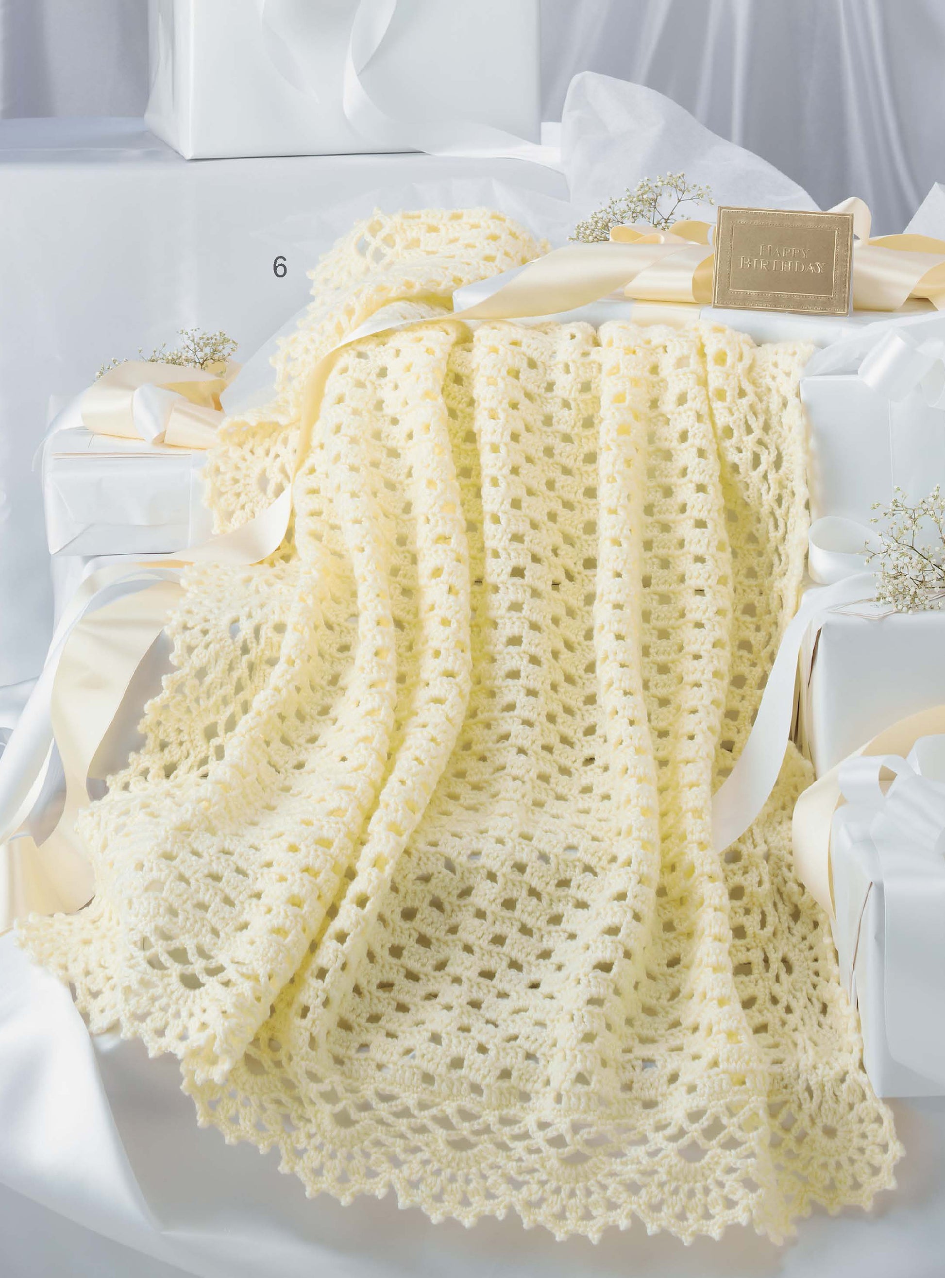The Keepsake Baby Afghans Digital Download by Leisure Arts features a soft, pale yellow crocheted blanket with elegant edgings and a lacy pattern, draped over white gift boxes decorated with wide ribbons and baby's breath.