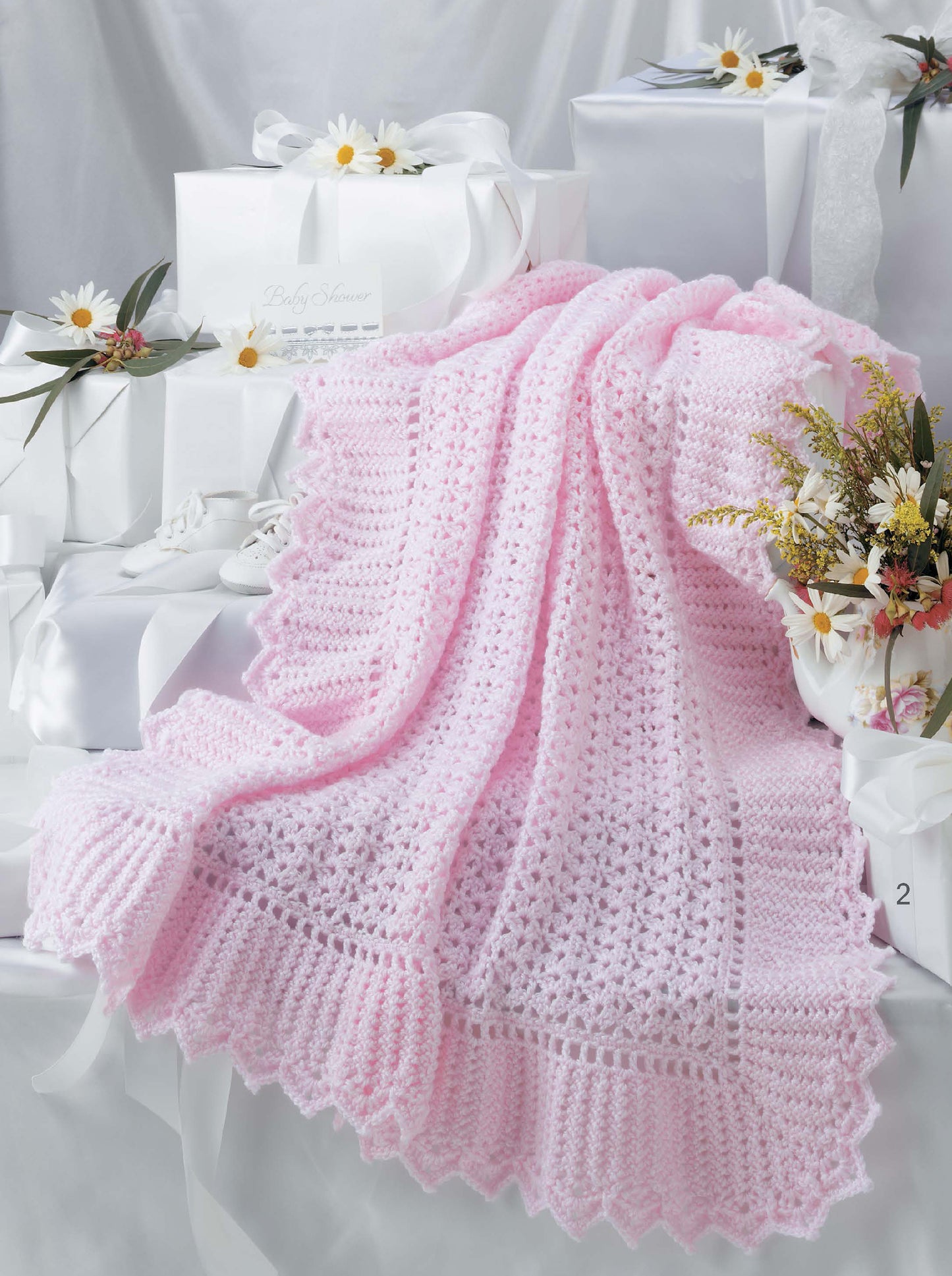 A soft pink crocheted blanket from the Keepsake Baby Afghans Digital Download by Leisure Arts is draped over white gift boxes with white ribbons and daisies, set against a light, elegant background.