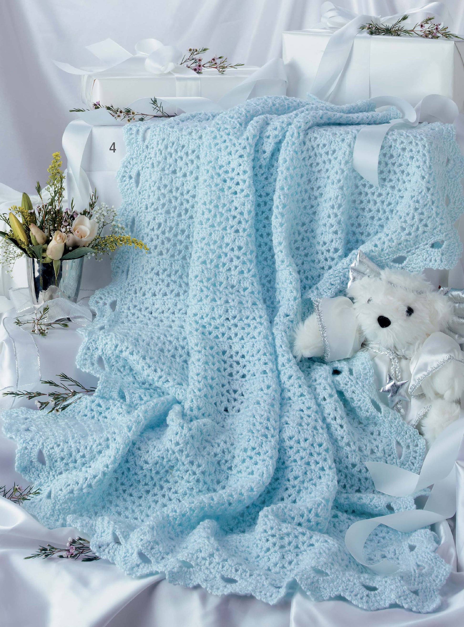 The Leisure Arts Keepsake Baby Afghans Digital Download features a light blue crocheted blanket with elegant edgings, draped over ribbon-tied white boxes, beside a white teddy bear and a small bouquet in a glass vase for old-fashioned charm.