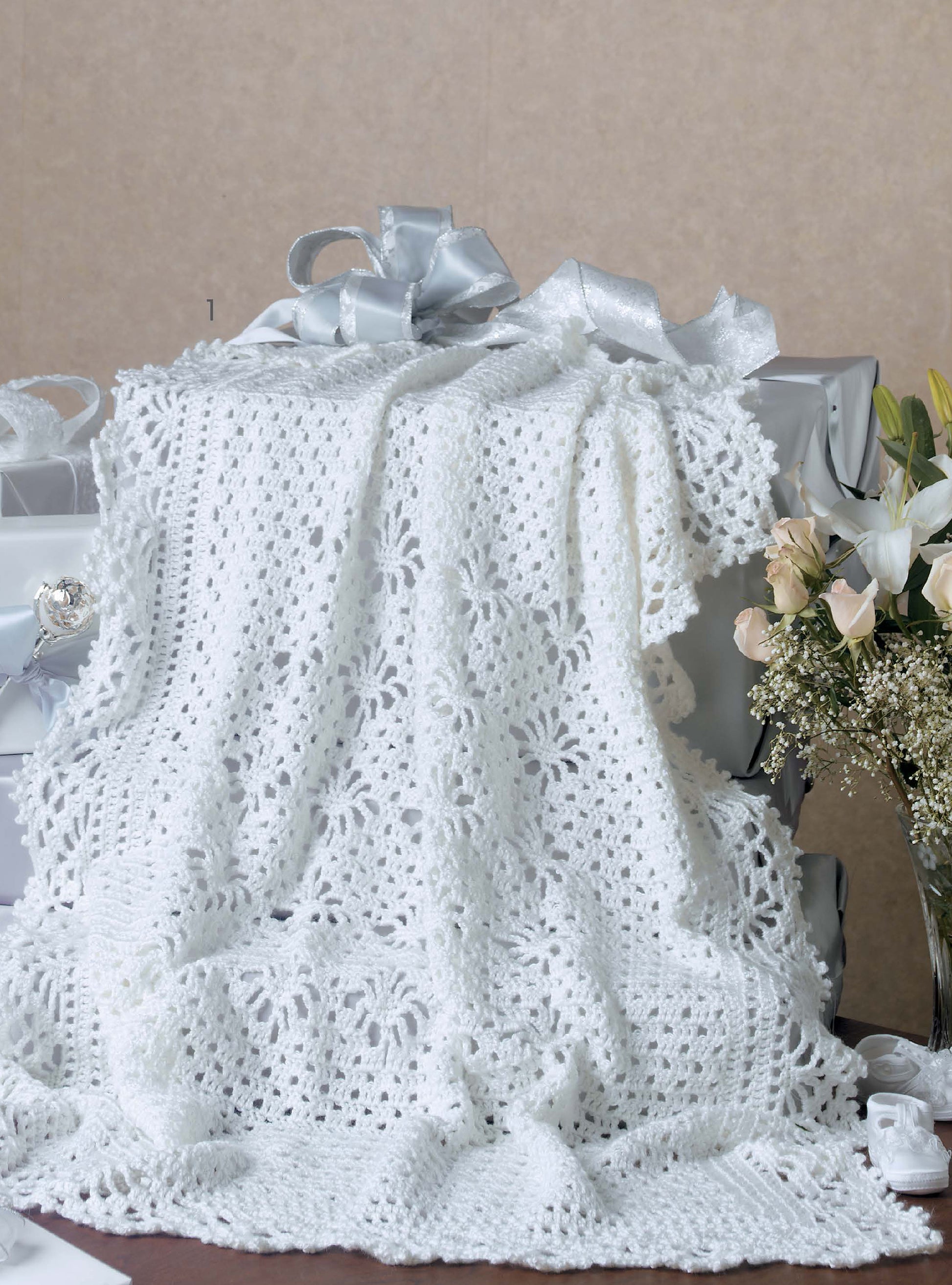 The Leisure Arts Keepsake Baby Afghans Digital Download features a white crocheted blanket with floral patterns and elegant edgings, adding old-fashioned charm to a table adorned with wrapped gifts and pale flowers.