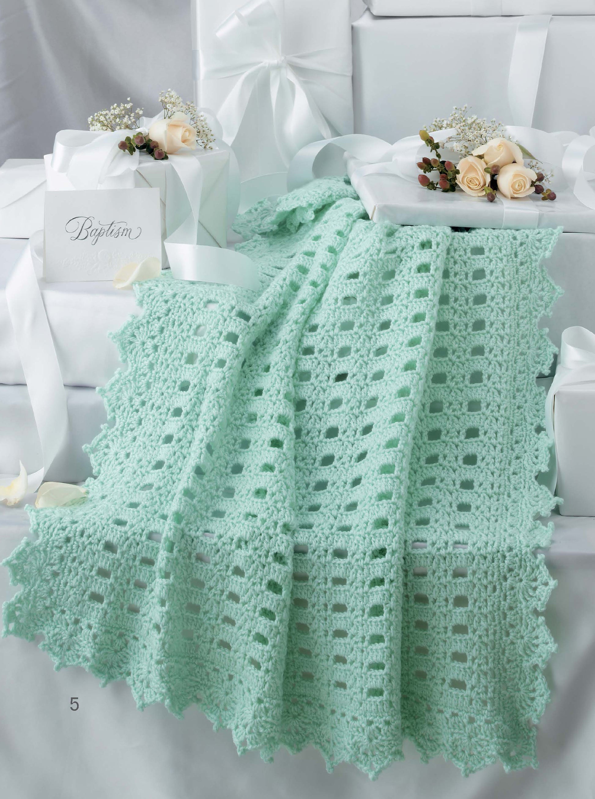 The Keepsake Baby Afghans Digital Download by Leisure Arts features a light green, lacy geometric blanket elegantly draped over white gift boxes adorned with white ribbons and small cream rose bouquets.