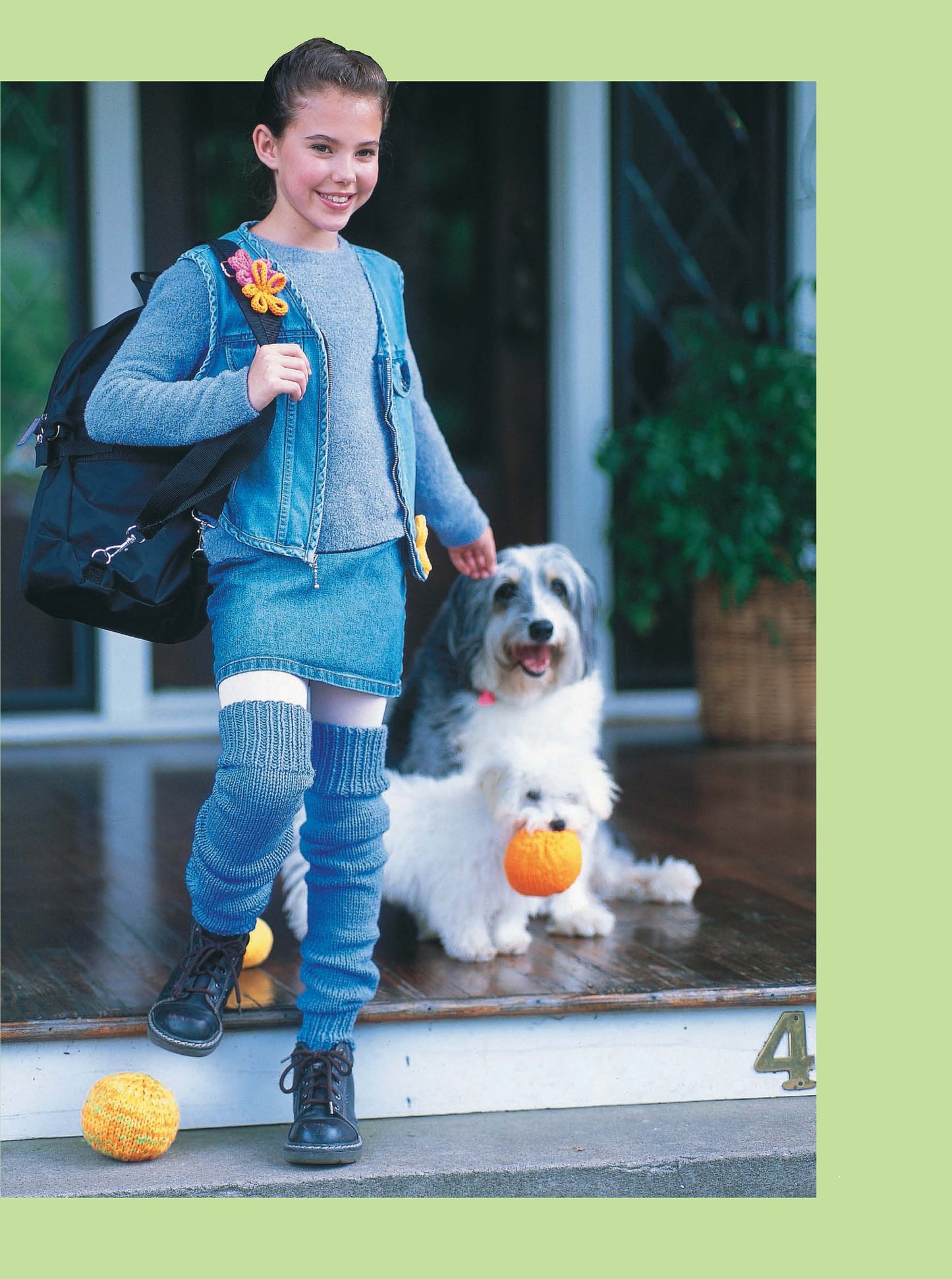 A happy girl in denim and blue leg warmers—made from the Leisure Arts Cool Stuff for Kids Teach Me to Knit digital download—stands with her backpack, while two fluffy dogs with orange balls sit beside her near a green house entrance.