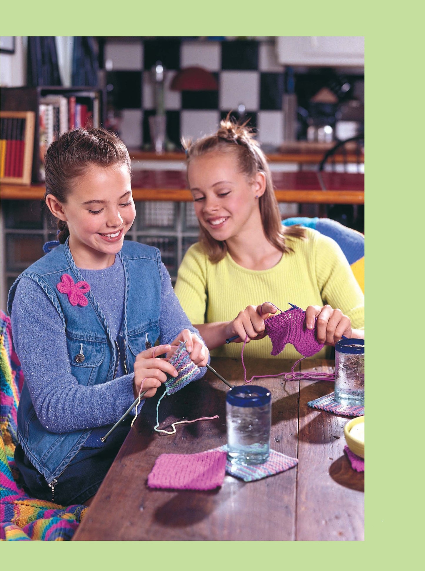 Two girls smile as they knit at a table with yarn and water glasses nearby. One wears a denim vest with a pink flower pin. The cozy room is perfect for using Leisure Arts' Cool Stuff for Kids Teach Me to Knit—12 Projects Digital Download.