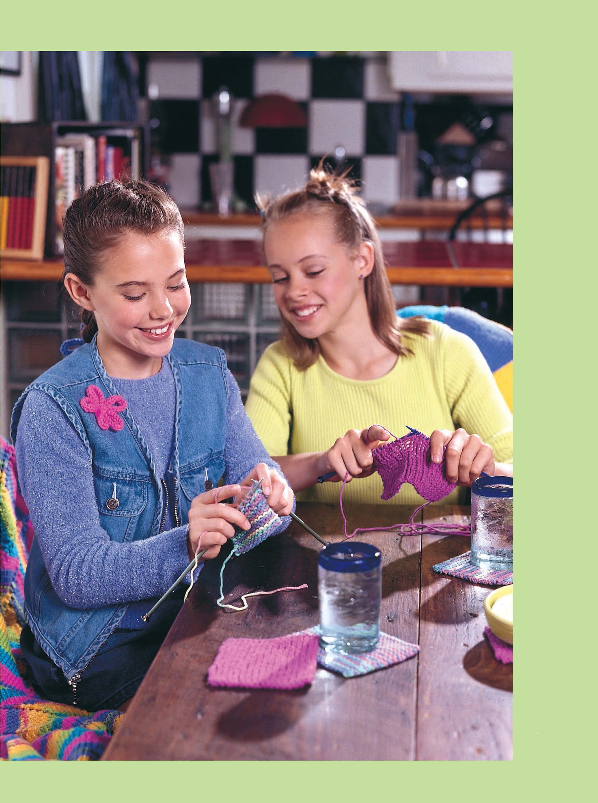 Two girls smile as they knit at a table with yarn and water glasses nearby. One wears a denim vest with a pink flower pin. The cozy room is perfect for using Leisure Arts' Cool Stuff for Kids Teach Me to Knit—12 Projects Digital Download.
