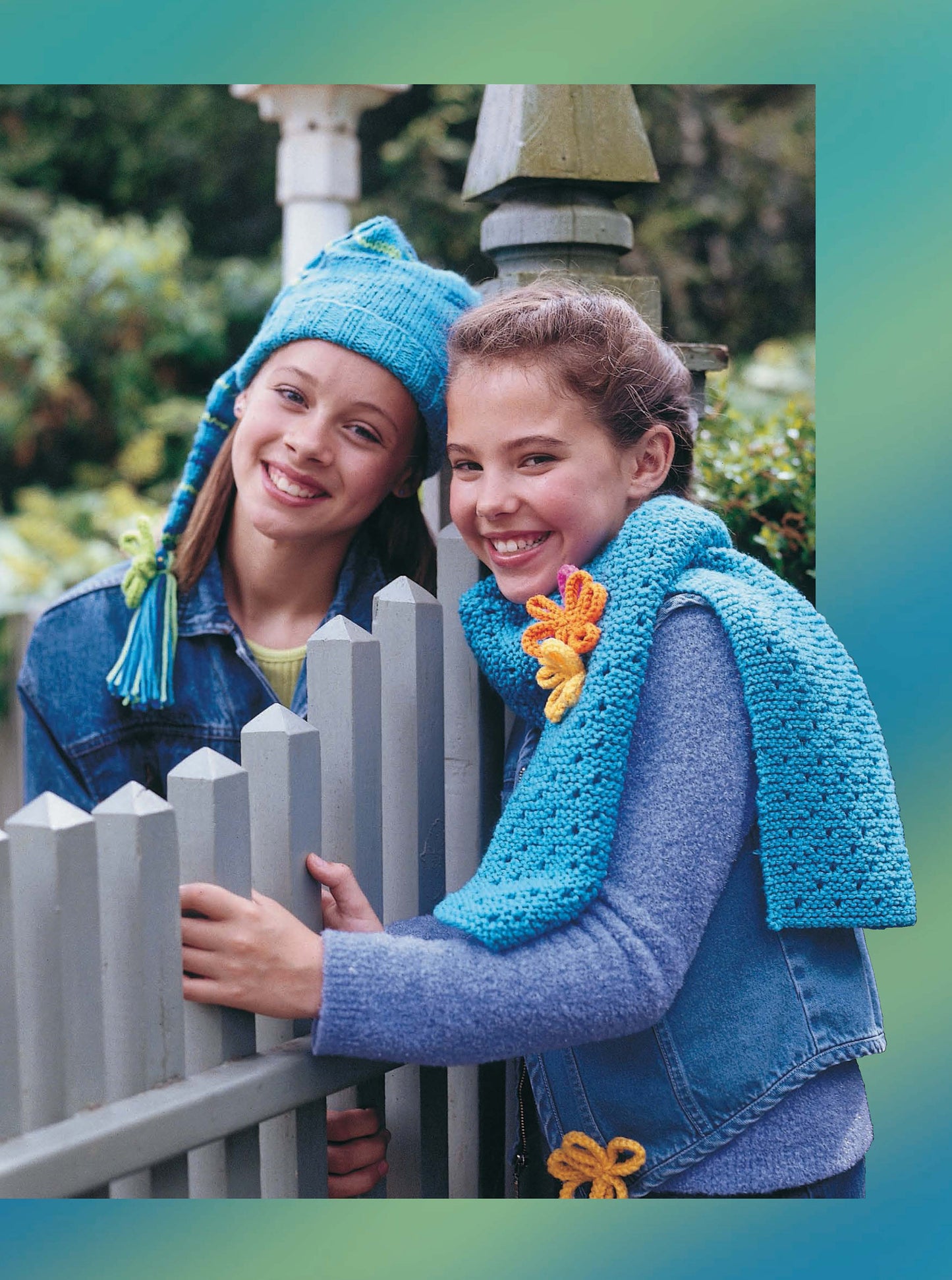 Two girls smile by a grey fence, showing off blue crochet accessories from Leisure Arts' "Cool Stuff for Kids Teach Me to Knit—12 Projects" digital download—perfect inspiration for young knitters. Green foliage frames the background.