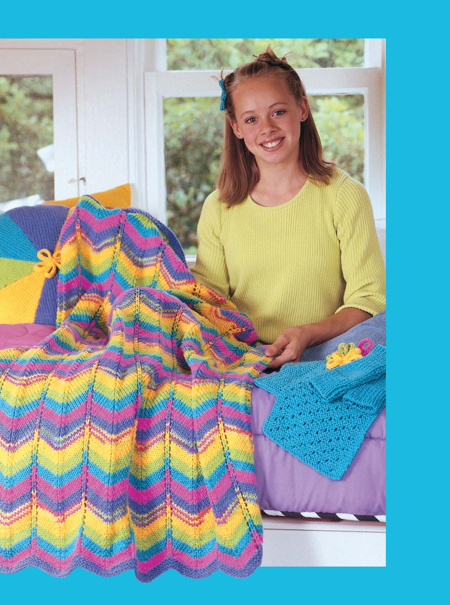 A smiling girl in a yellow sweater sits on a bed, holding a colorful knit blanket made from "Cool Stuff for Kids Teach Me to Knit—12 Projects Digital Download" by Leisure Arts, with more blue and purple knits beside her.