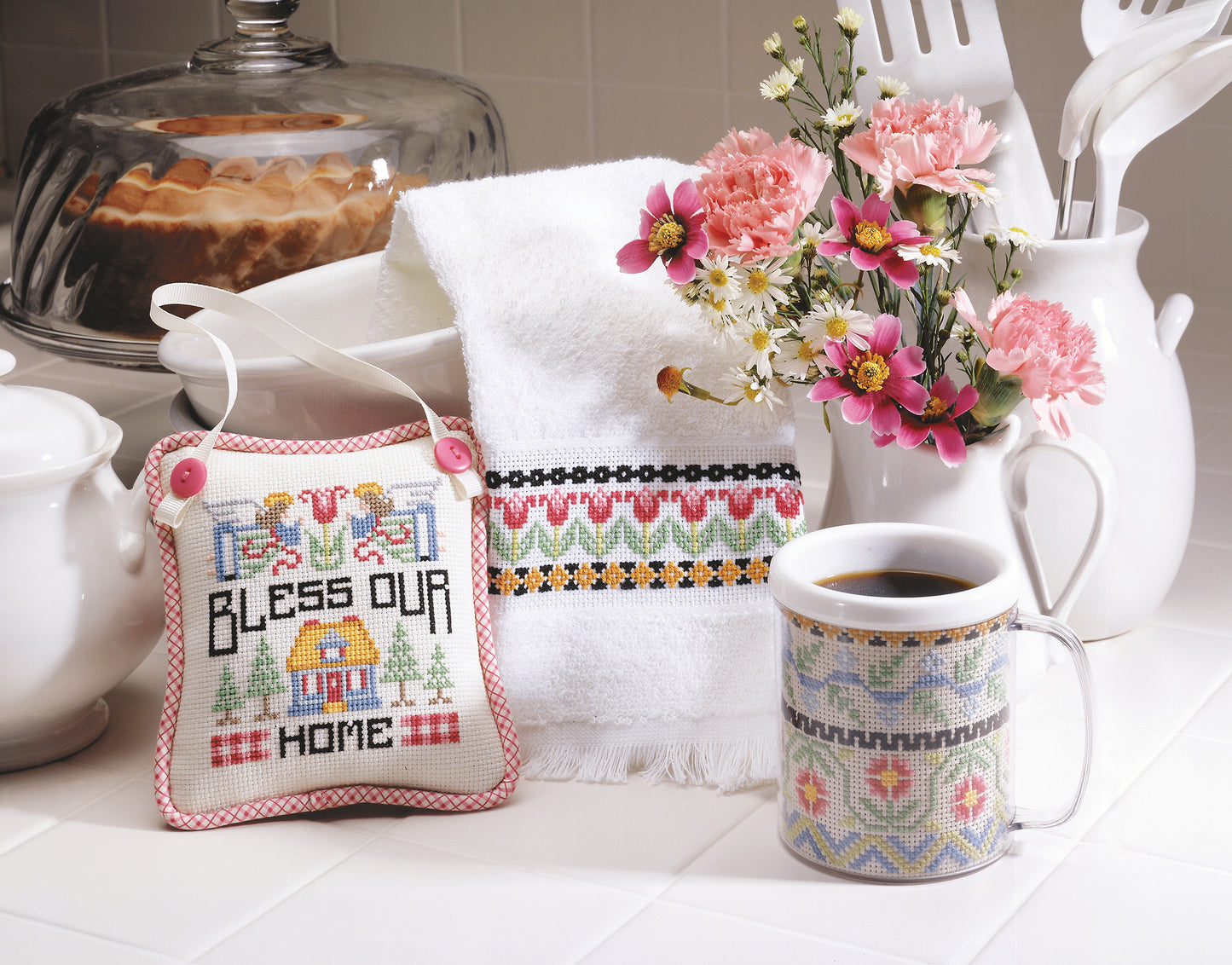 A cozy kitchen scene featuring Leisure Arts' "A Sampling of Tradition" cross stitch (digital download), embroidered home accents, a mug with an embroidered sleeve, and pink and white flowers on a white countertop.