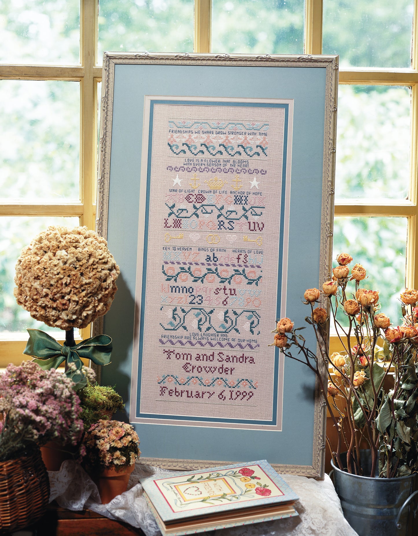 The "A Sampling of Tradition Cross Stitch Digital Download" by Leisure Arts is displayed in a frame on a sunlit table, surrounded by dried flowers and potted plants, its pastel patterns and text enhanced by the natural light.