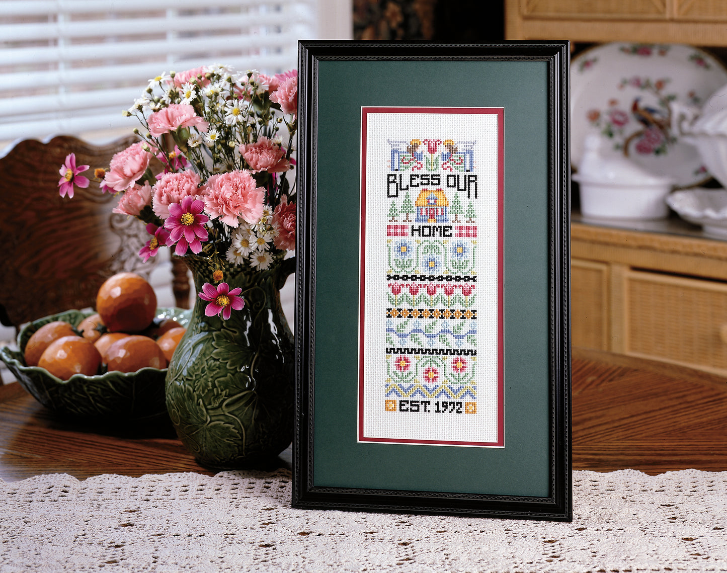 A Sampling of Tradition Cross Stitch Digital Download by Leisure Arts is displayed in a frame on a lace tablecloth beside pink flowers and oranges, bringing cozy charm to a bright dining room.