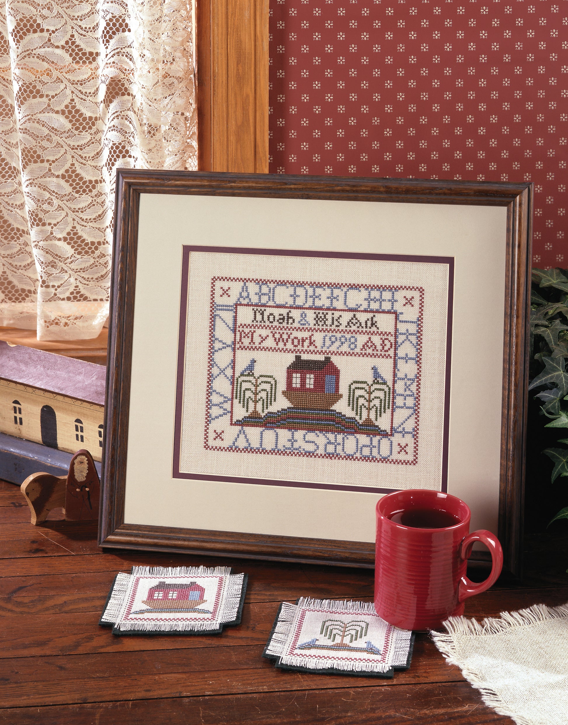 A Sampling of Tradition Cross Stitch Digital Download by Leisure Arts features Noah's Ark artwork. Displayed on a wooden table by a red mug, matching coasters, and a lace curtain—perfect for cross stitch wall hanging enthusiasts.