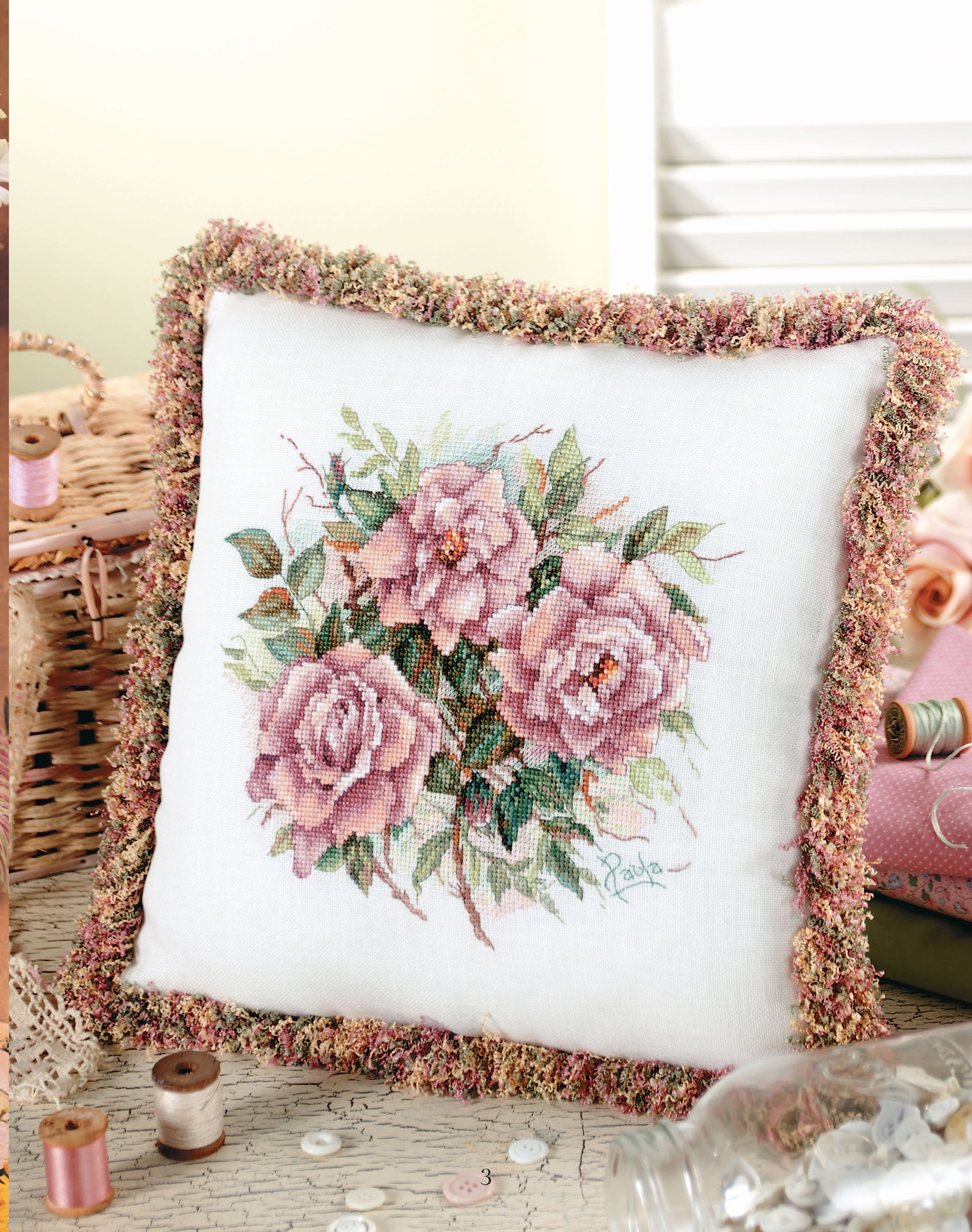 The "Pillows from Paula's Garden" digital download by Leisure Arts features a decorative pillow with pink roses and green leaves, trimmed in multicolors and styled among sewing supplies like thread, buttons, and a wicker basket.