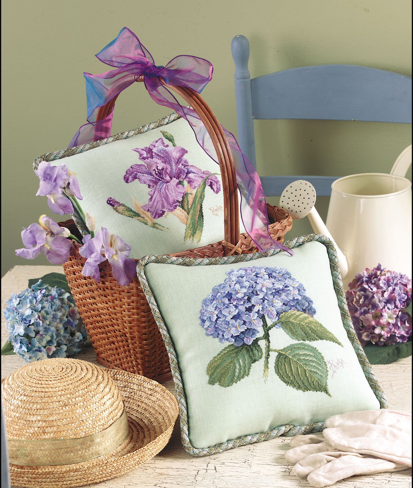 Leisure Arts' Pillows from Paula's Garden Digital Download, a straw hat, gardening gloves, a watering can, and a woven basket with purple ribbon are displayed on a rustic table beside fresh flowers and a blue chair.