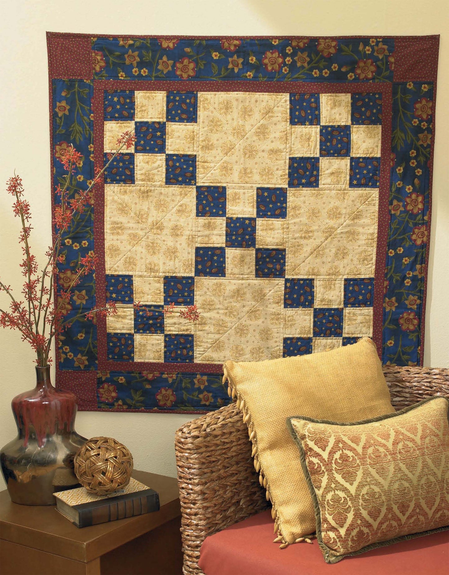 The "I Can't Believe I'm Quilting" digital download by Leisure Arts is ideal for beginner quilters and features blue, tan, and red patchwork. Shown above a wicker chair with gold pillows and next to a brown vase with red branches.