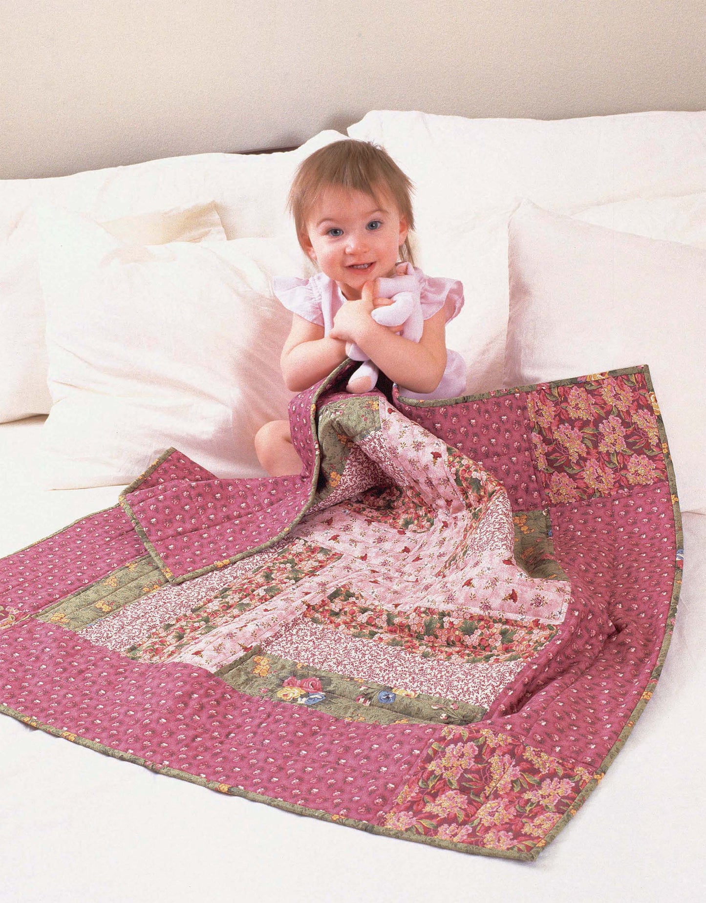 A smiling toddler hugs a stuffed animal on a bed with a pink floral patchwork quilt, inspired by beginner projects from the "I Can't Believe I'm Quilting Digital Download" by Leisure Arts. White pillows and bedding complete the cozy scene.