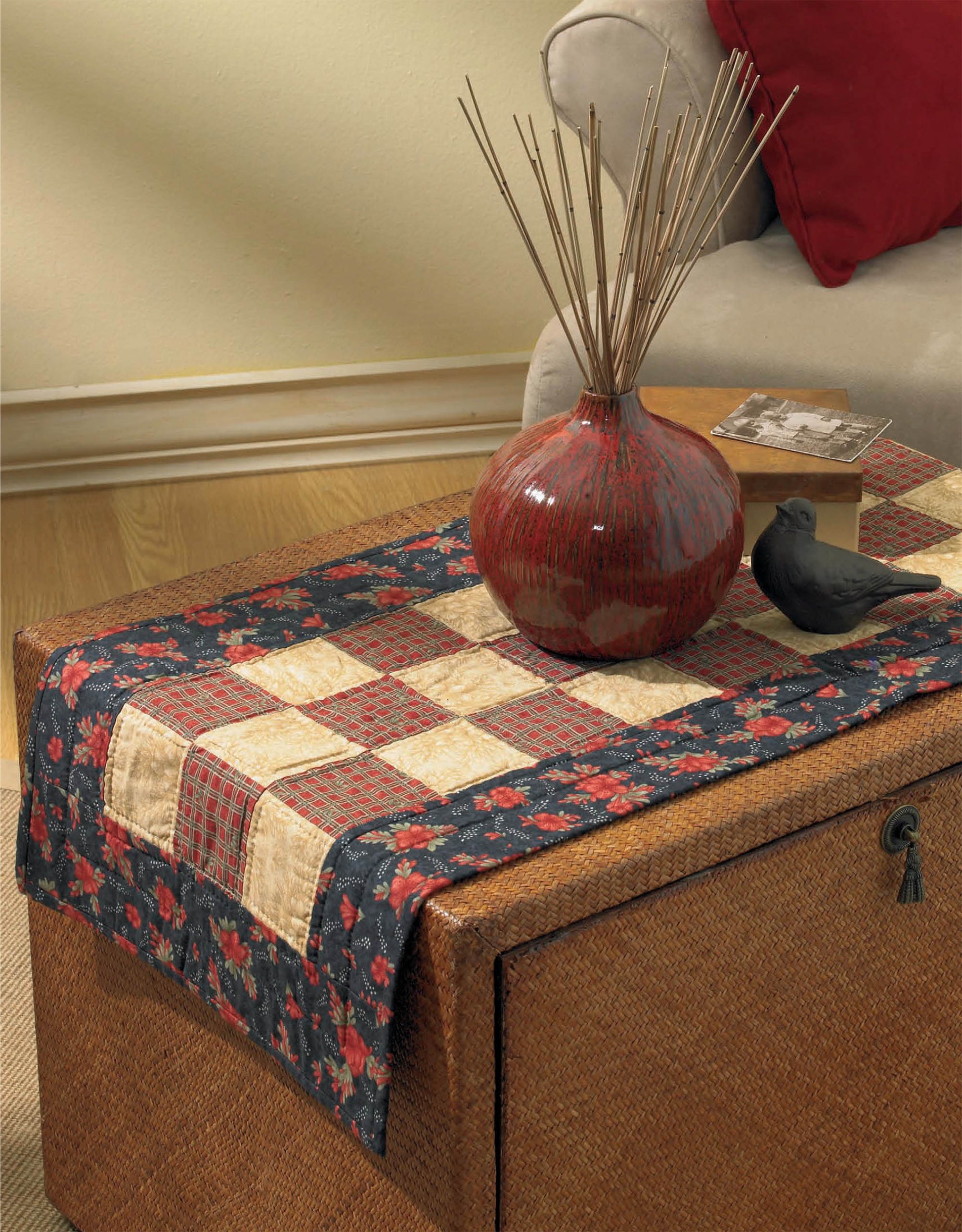 A wicker storage chest topped with a table runner made from Leisure Arts’ I Can’t Believe I’m Quilting Digital Download displays a red ceramic vase with reed sticks, a decorative box, and a black bird figurine beside a beige sofa with a red pillow.