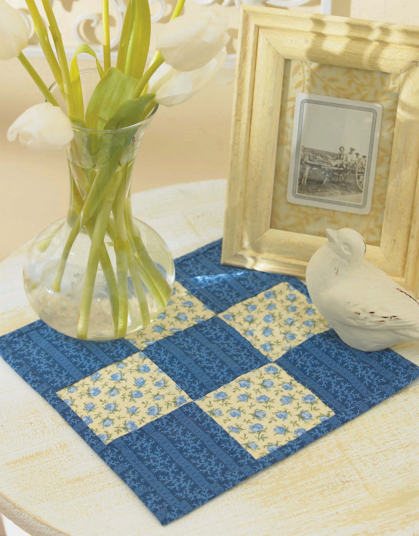 A clear vase of white tulips, a framed black-and-white photo, and a white ceramic bird sit on a table with a blue and yellow patchwork mat inspired by Leisure Arts' "I Can't Believe I'm Quilting" digital download.