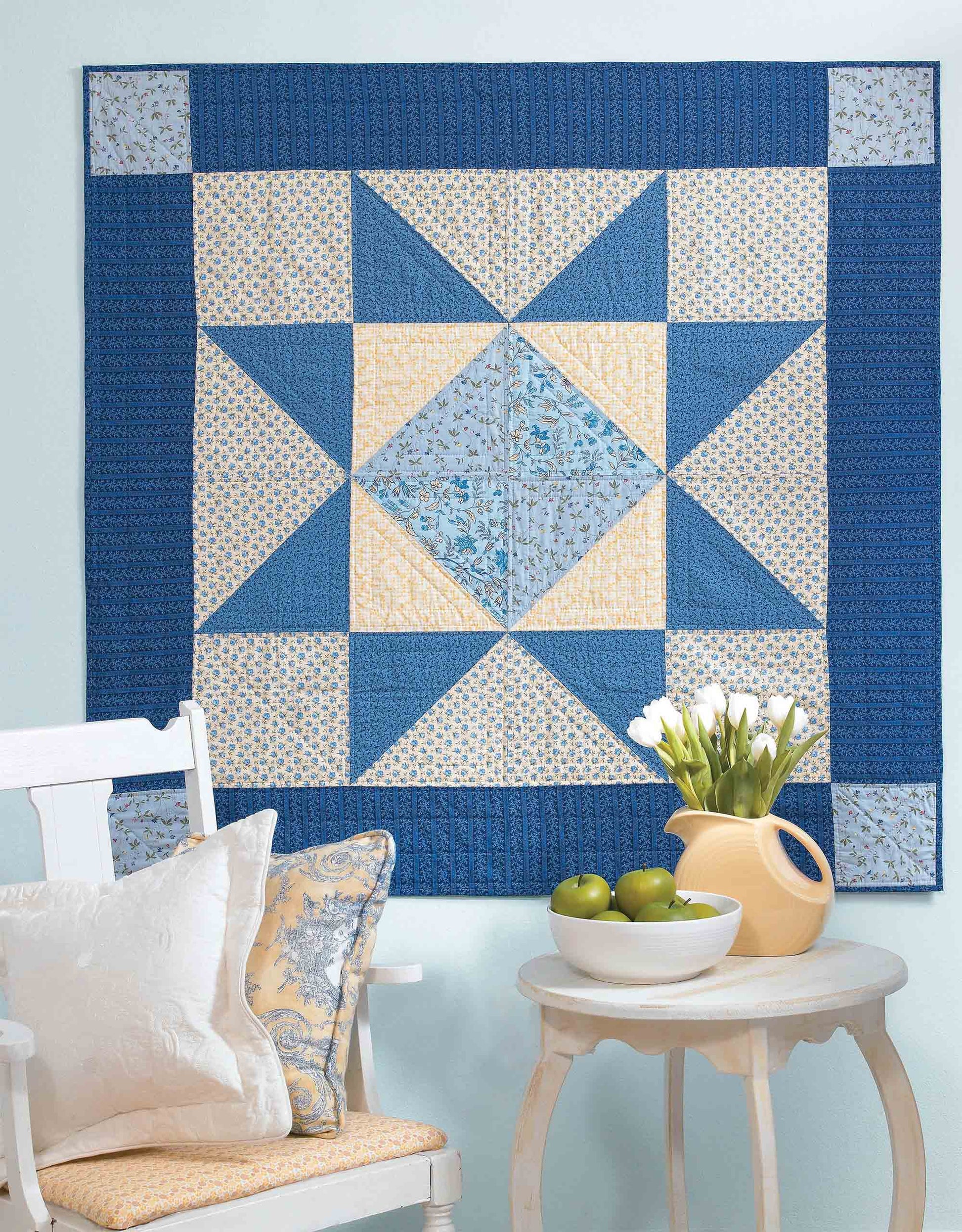 The "I Can't Believe I'm Quilting" digital download by Leisure Arts is showcased on a blue wall above a white chair with patterned pillows and a table featuring green apples, a yellow pitcher, and white flowers in a vase.