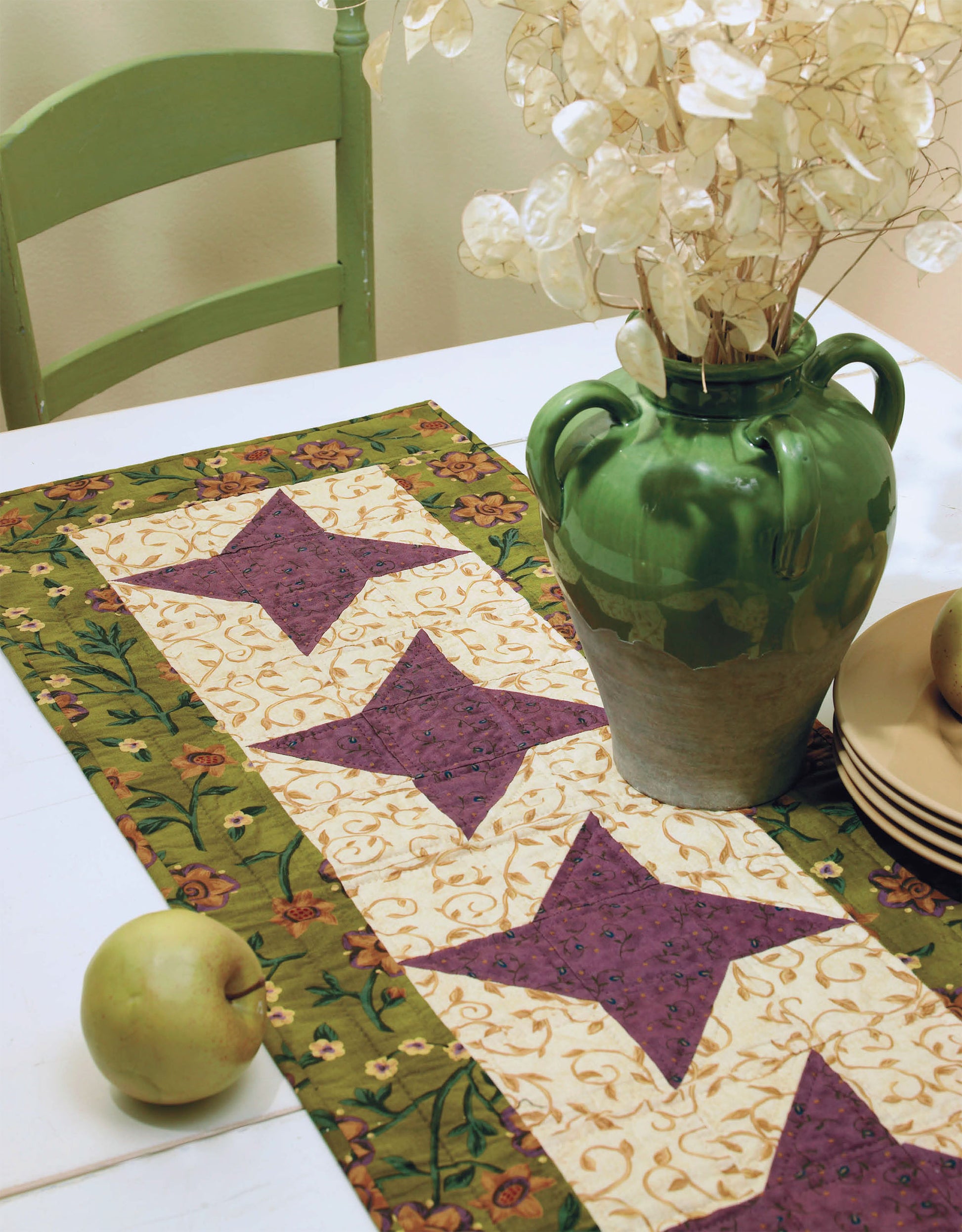 The "I Can't Believe I'm Quilting Digital Download" by Leisure Arts features a purple and cream star-patterned table runner, perfect for beginner quilting. Displayed on a white table with plates, apples, a green chair, and a vase of dried flowers.