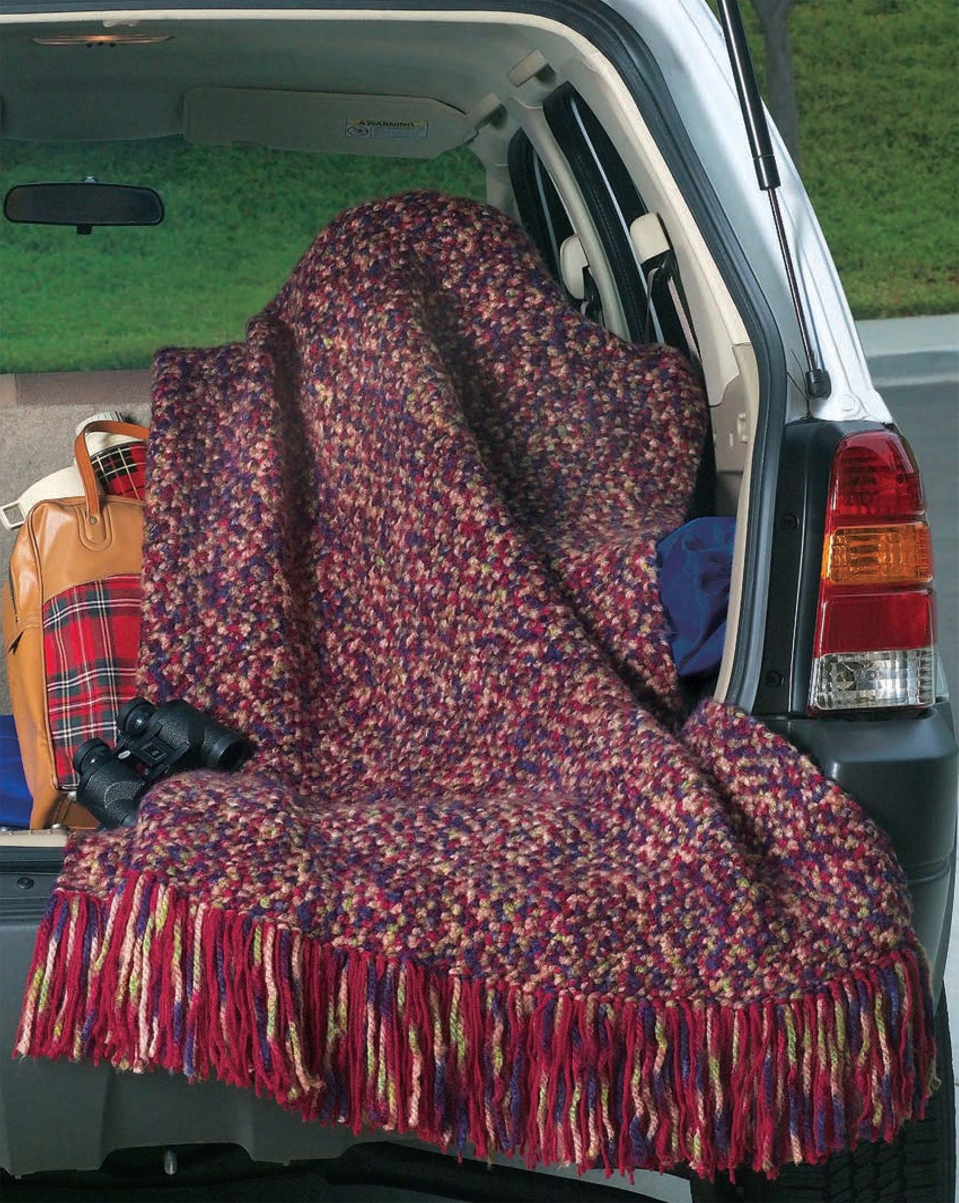 A vibrant knit blanket inspired by Leisure Arts' 48-Hour Afghans Digital Download drapes over an SUV's open back. Nearby are a plaid bag and binoculars, with grass and pavement visible in the background. Perfect for new crochet pattern ideas.
