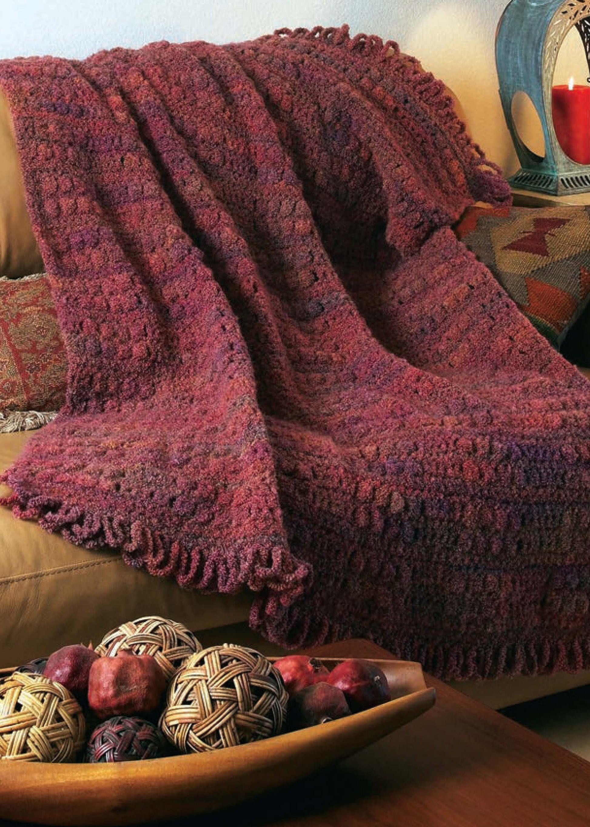 A burgundy crochet blanket made with classic patterns from the 48-Hour Afghans Digital Download by Leisure Arts is draped over a tan couch, with a wooden bowl of twine and fabric balls in natural and red tones in the foreground.