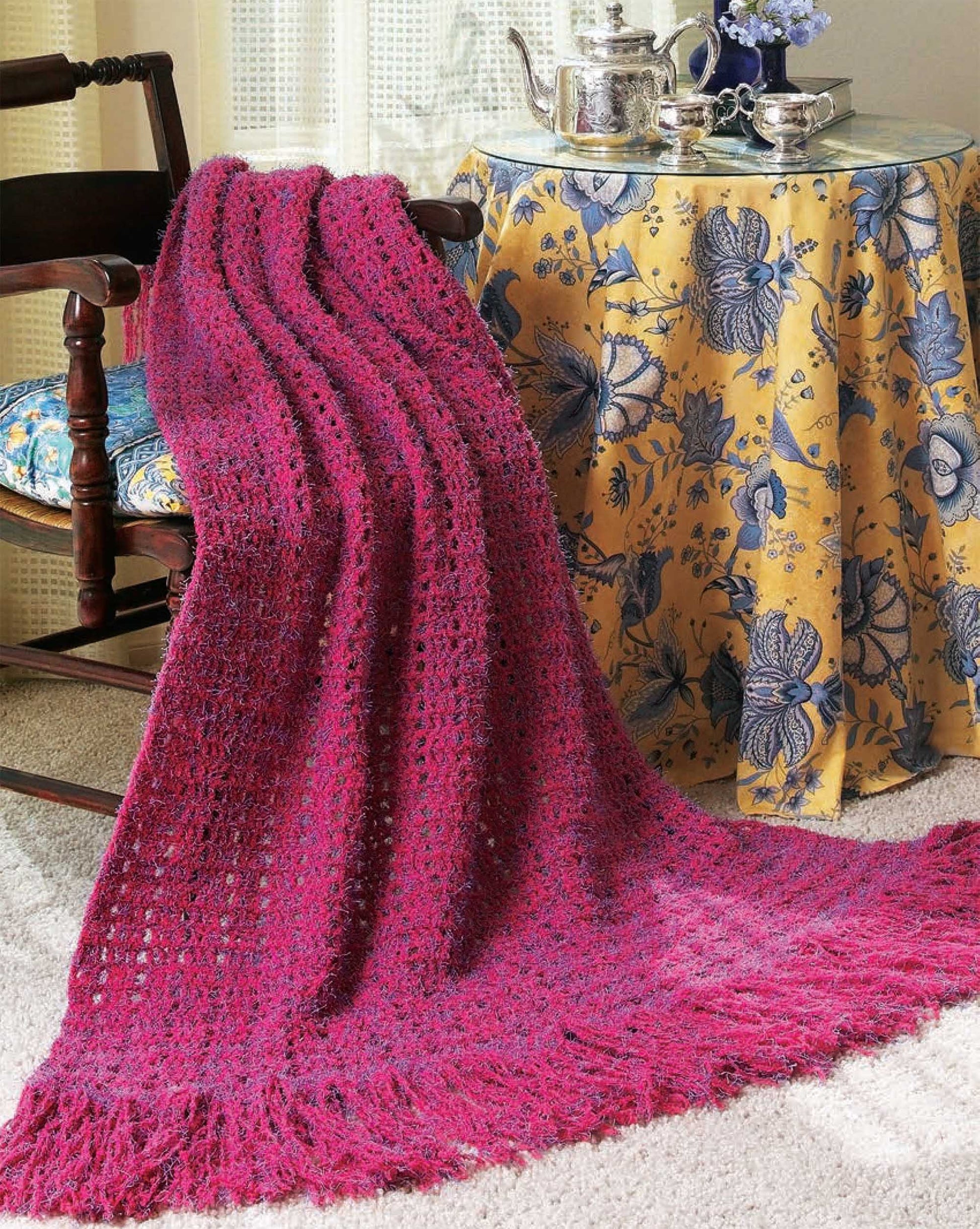 A vibrant pink crochet blanket from Leisure Arts’ 48-Hour Afghans Digital Download drapes over a chair beside a table set with a floral cloth, teapot, and vase—perfect for displaying beautiful afghan patterns in your home.